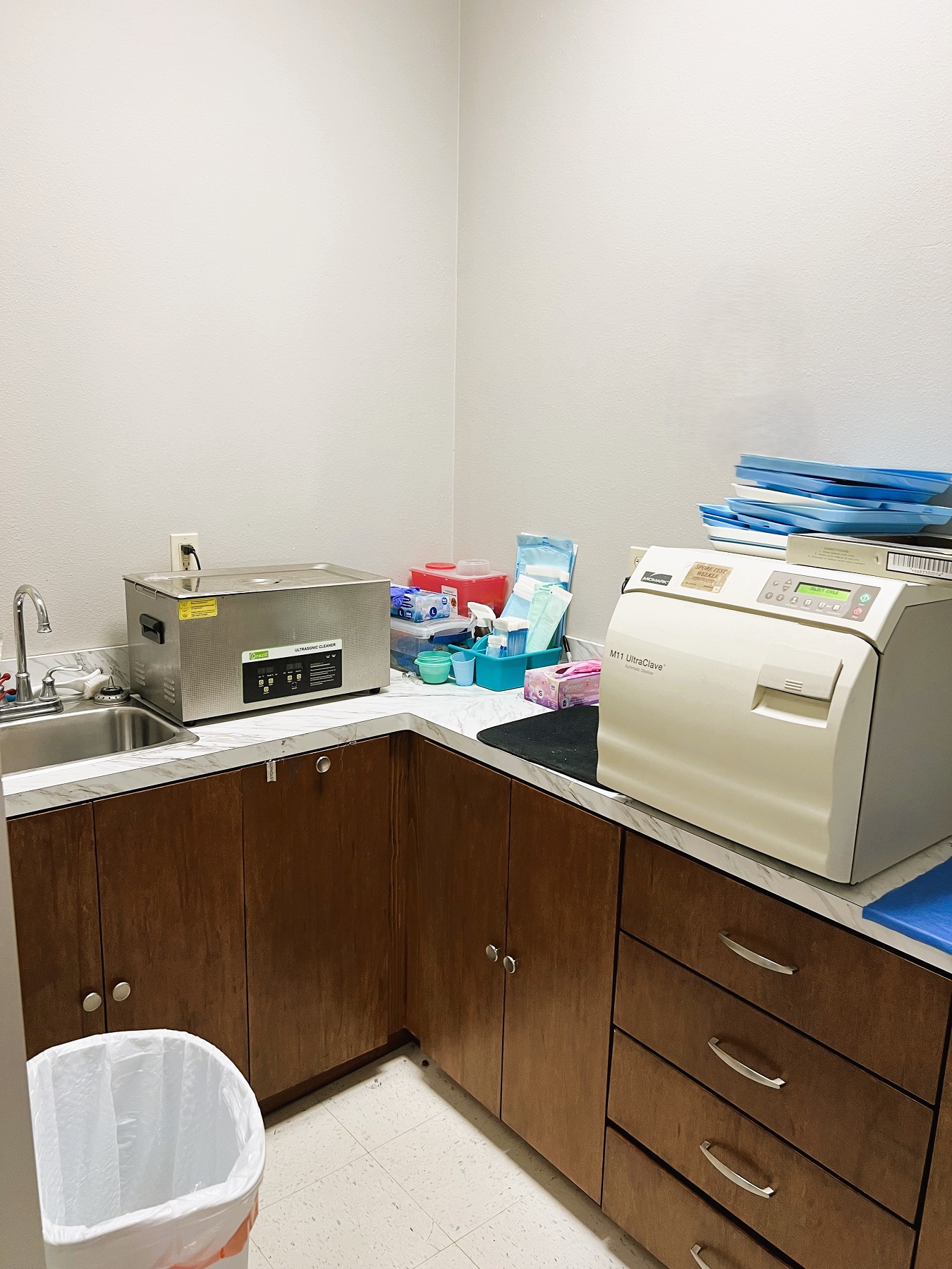 Light colored walls with dark baseboards and white, large, tile flooring. Dark stained cabinetry with light counter tops holds lots of dental sterilization tools, equipment, and supplies.