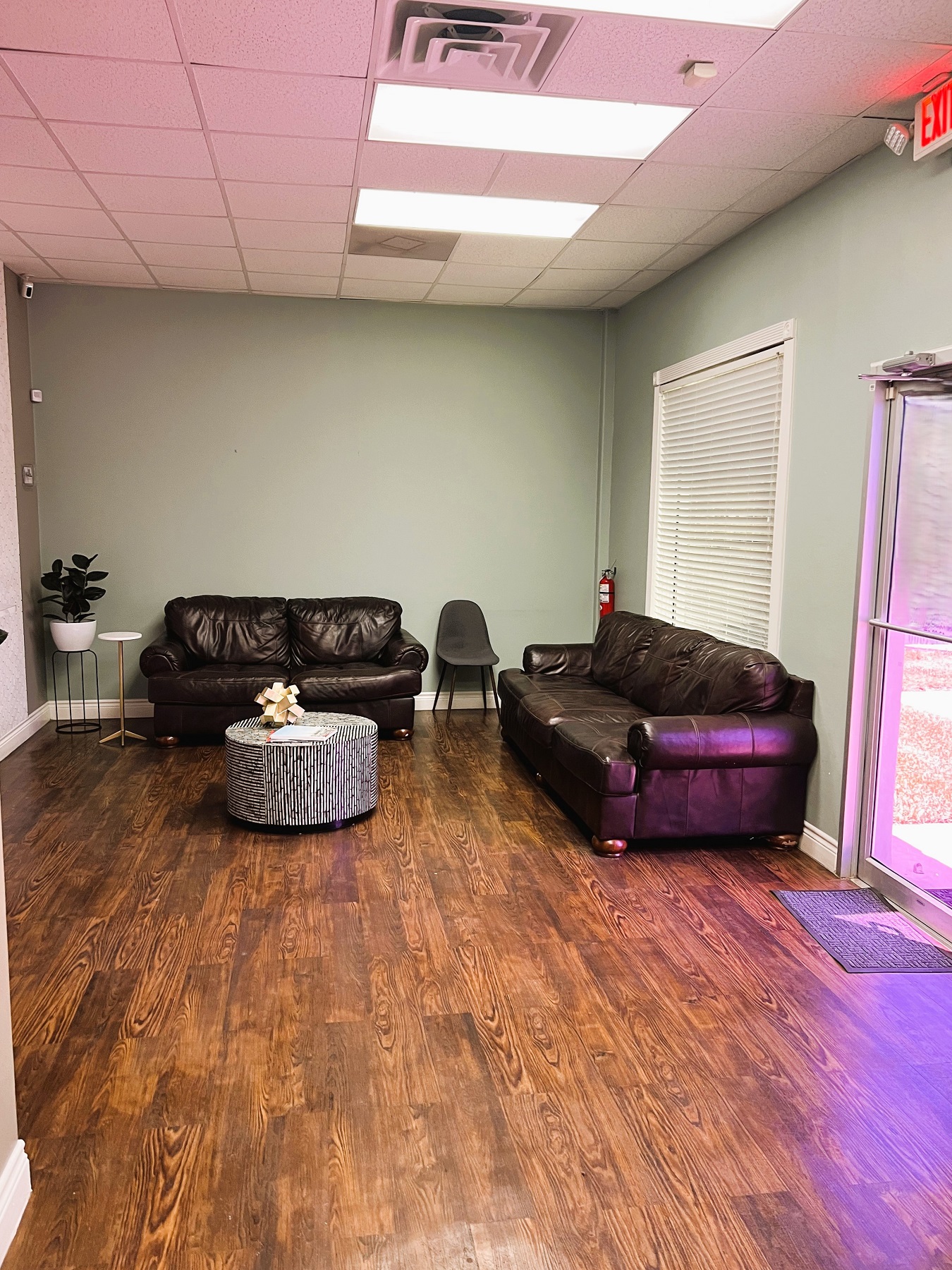 Light colored walls with white baseboards and trim. Wood-look flooring. Overstuffed couches and a chair surround a round coffee table. A large window next to the entry door to the practice.