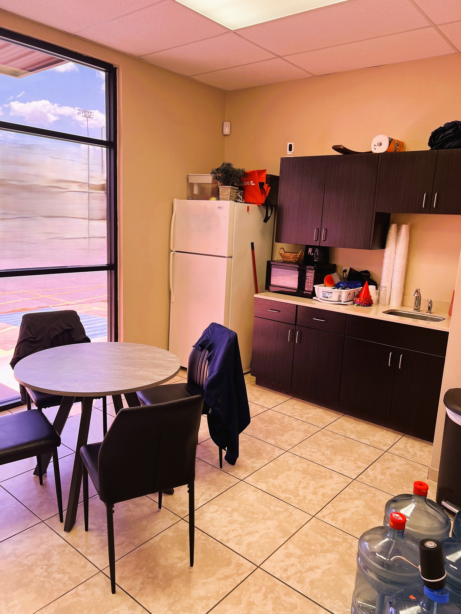 Light colored walls with floor to ceiling store front windows. A white fridge sits in the corner, black cabintery with tan counter tops and a small stainless steel sink. Large tan tile flooring with matching baseboards. Round table is surrounded by black cushioned chairs