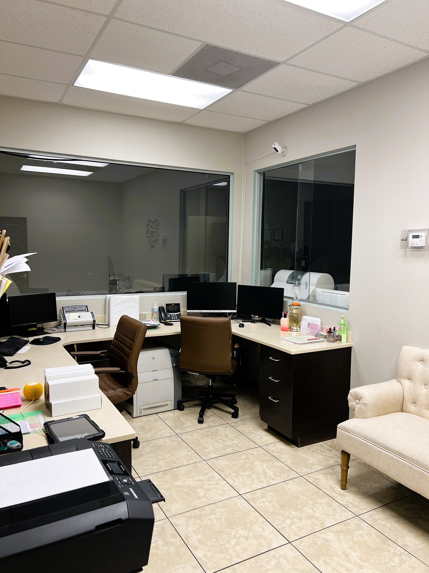Light colored walls with huge windows. "U" shaped built in desk with a tan top and black rolling office chairs holds lots of office equipment and supplies. An overstuffed couch sits at the end of the desk against the walls.