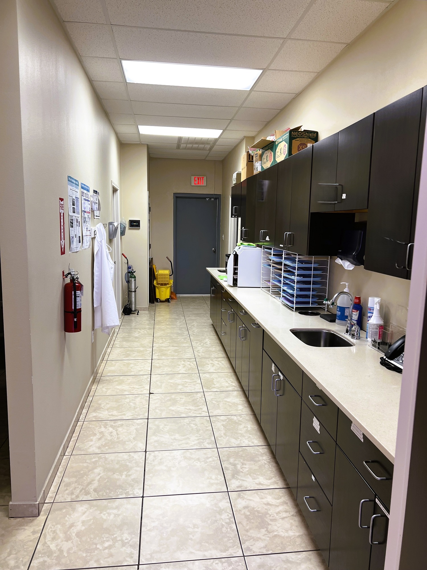 Light colored walls with large tan tile flooring and matching baseboards. Black cabinetry stretches along the whole right hand wall with tan countertops. This holds lots of dental lab tools, equipment, and supplies.