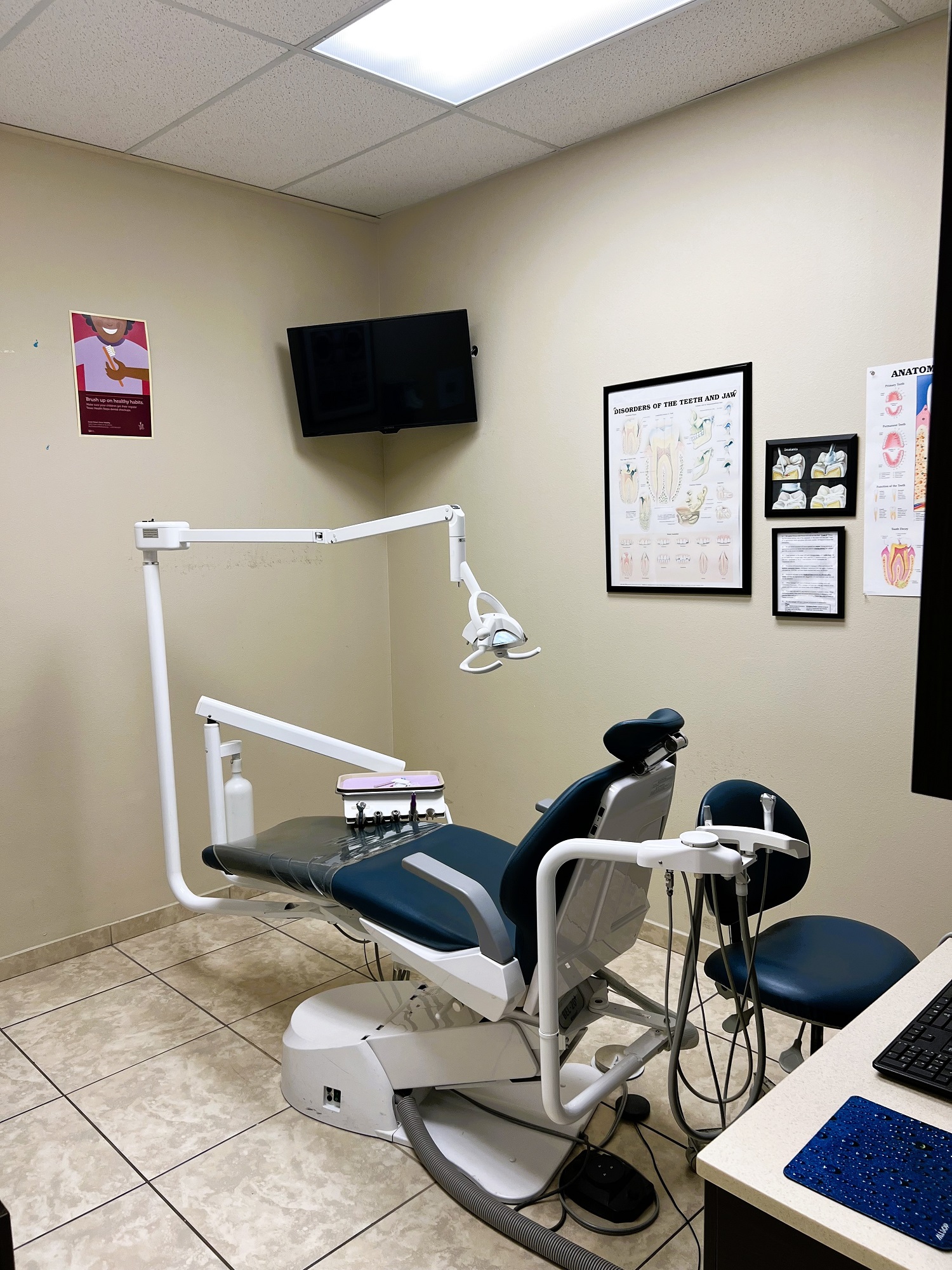 Dental operatory with dark dental procedural chair anchoring the room. Light colored walls with large square floor tile and matching baseboard. Dental procedural light over the chair with rolling dr chair. Flat screen tv mounted high in the corner. Dark cabinet with tan counter-top holds lots of dental tools, supplies, and equipment.