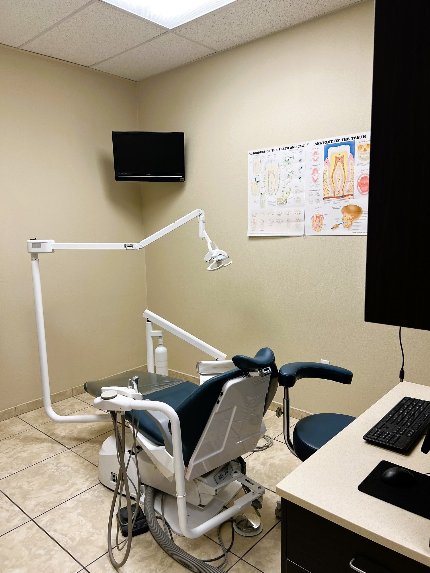 Dental operatory with dark dental procedural chair anchoring the room. Light colored walls with large square floor tile and matching baseboard. Dental procedural light over the chair with rolling dr chair. Flat screen tv mounted high in the corner. Dark cabinet with tan counter-top holds lots of dental tools, supplies, and equipment.