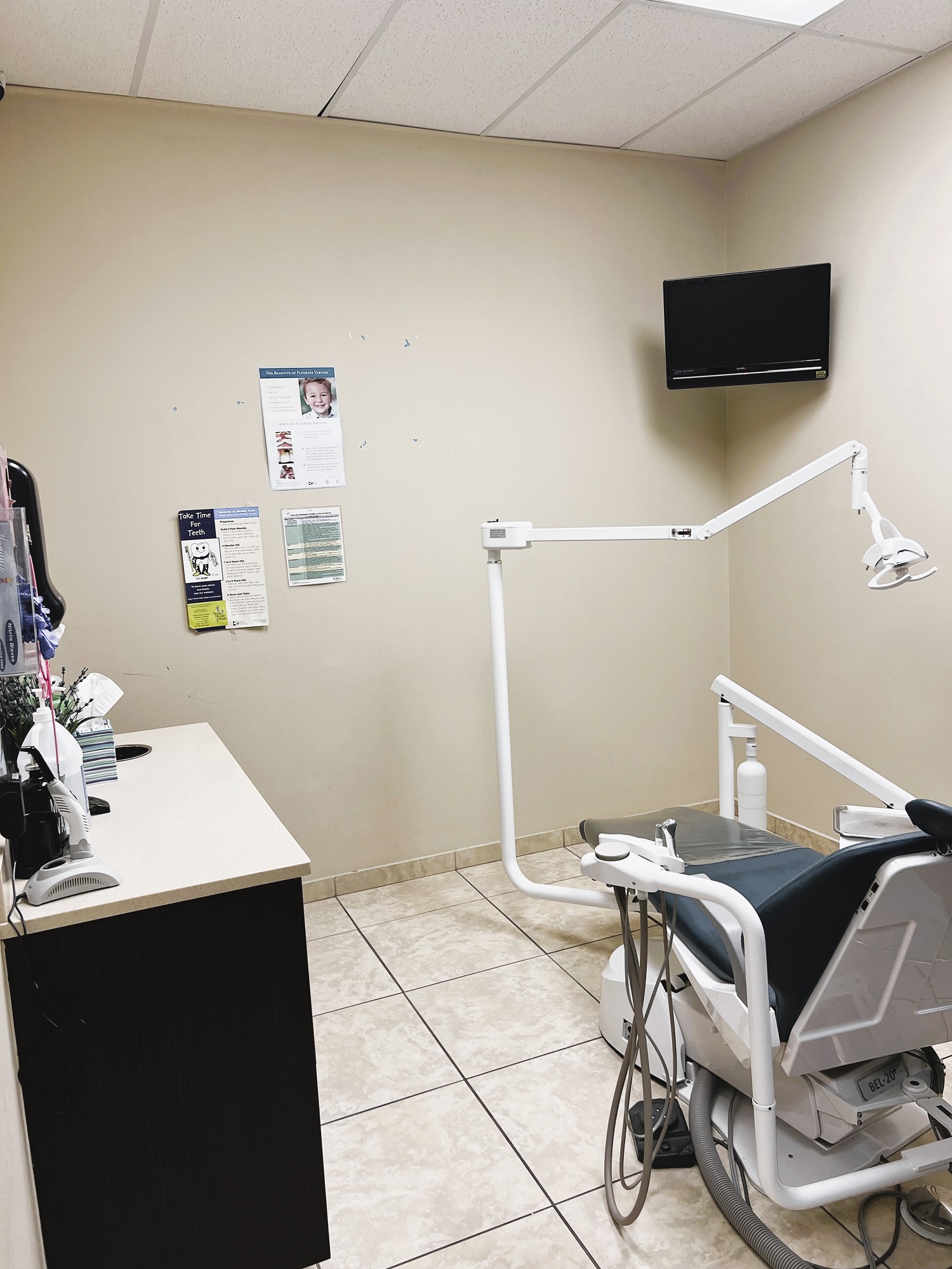 Dental operatory with dark dental procedural chair anchoring the room. Light colored walls with large square floor tile and matching baseboard. Dental procedural light over the chair with rolling dr chair. Flat screen tv mounted high in the corner. Dark cabinet with tan counter-top holds lots of dental tools, supplies, and equipment.