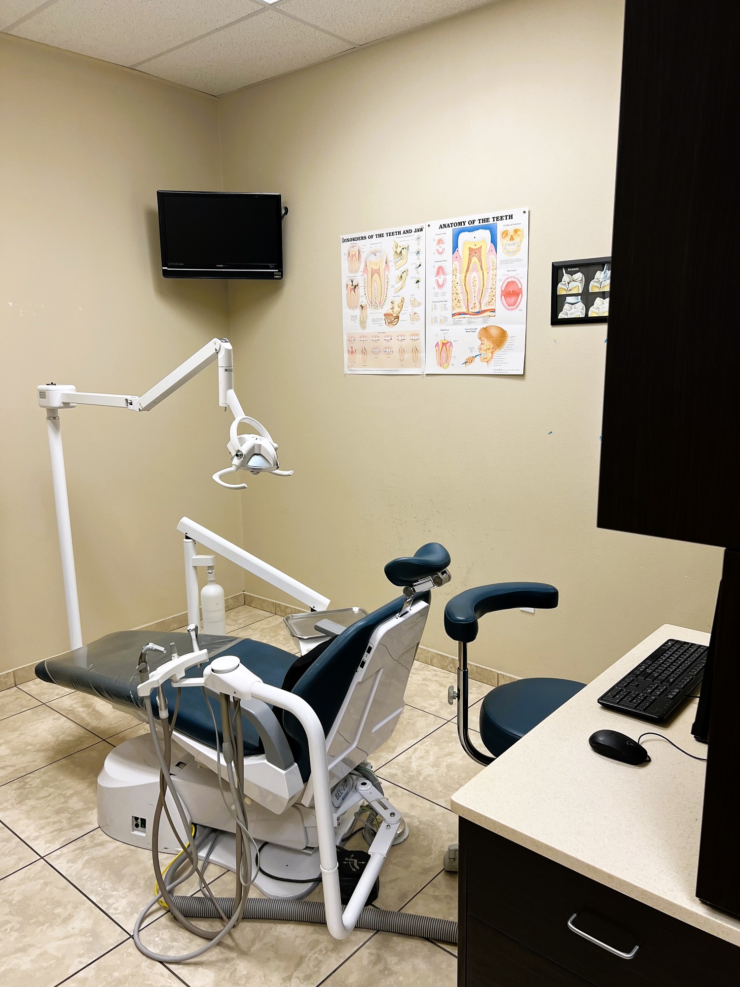 Dental operatory with dark dental procedural chair anchoring the room. Light colored walls with large square floor tile and matching baseboard. Dental procedural light over the chair with rolling dr chair. Flat screen tv mounted high in the corner. Dark cabinet with tan counter-top holds lots of dental tools, supplies, and equipment.