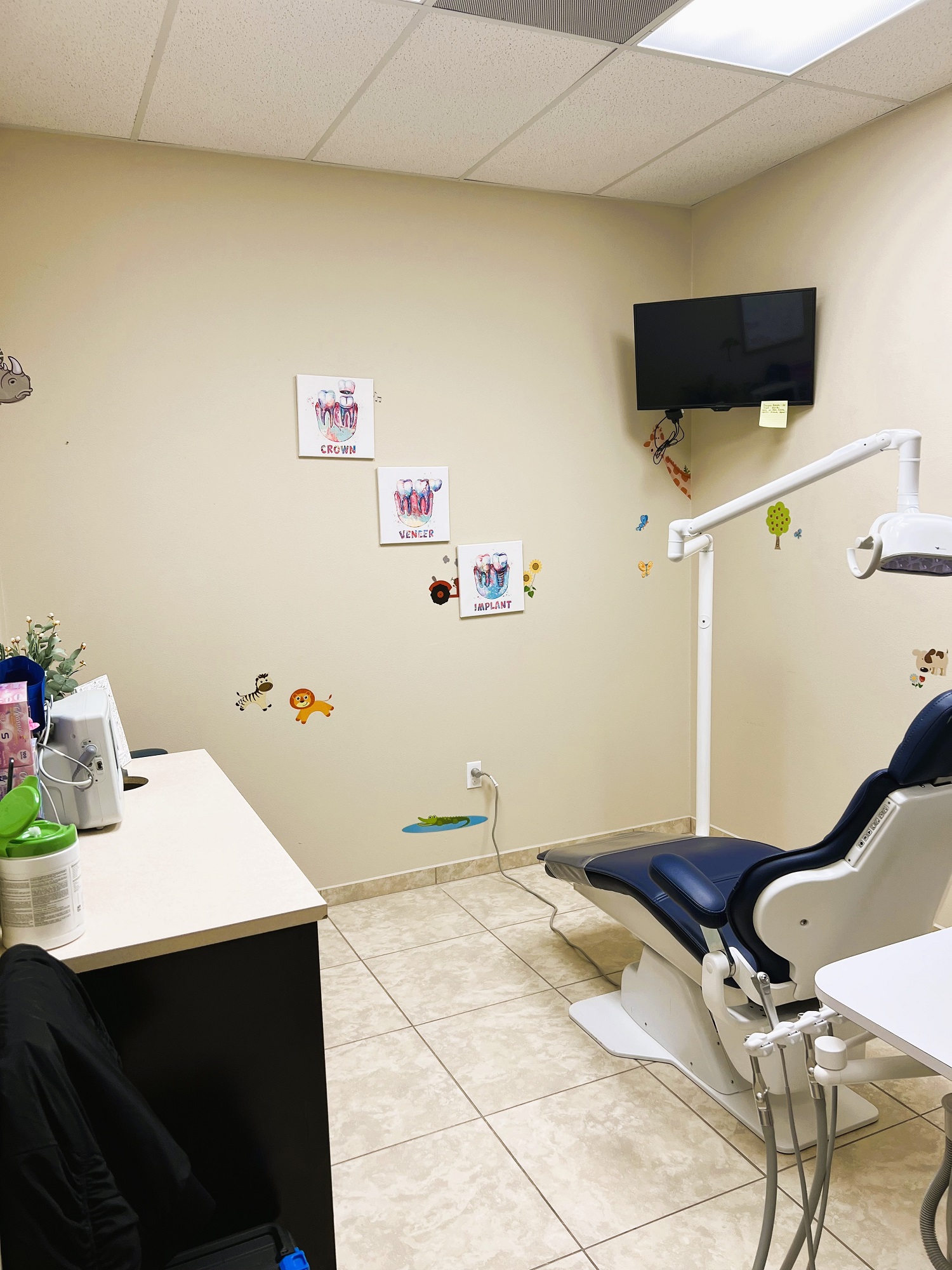 Dental operatory with dark dental procedural chair anchoring the room. Light colored walls with large square floor tile and matching baseboard. Dental procedural light over the chair with rolling dr chair. Flat screen tv mounted high in the corner. Dark cabinet with tan counter-top holds lots of dental tools, supplies, and equipment.