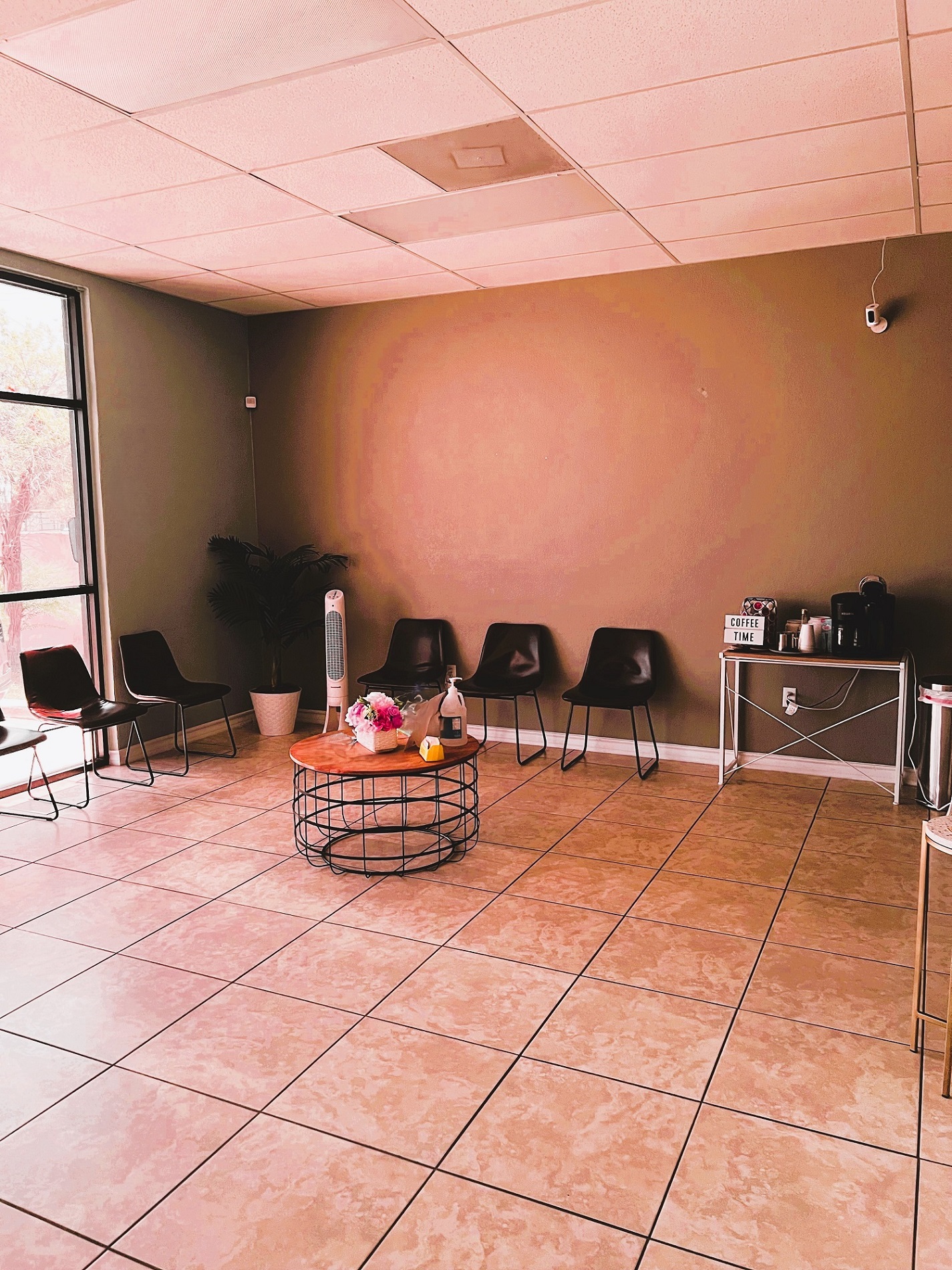 Colored accent wall with store front windows on the front of the practice. Large, square tile in the waiting area. Cushioned chairs line both walls with a large plant in the corner and a round coffee table in the center. There's a small end table with a little coffee bar at the end of the chairs.