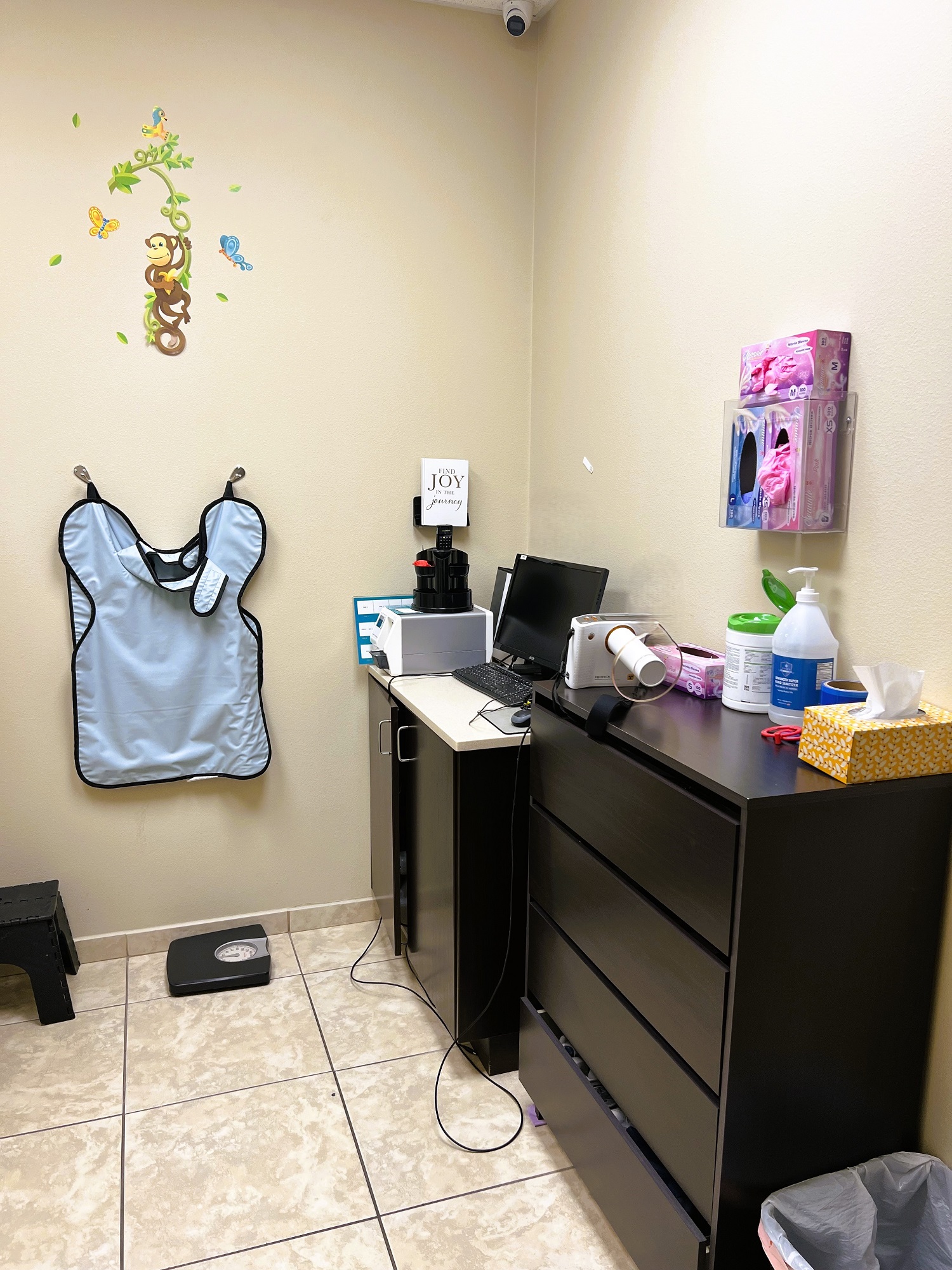 Light colored walls with large tan tile flooring with matching baseboard. Lead apron hangs on the wall. Black cabinetry sits against the wall with a tan countertop holds lots of dental equipment.