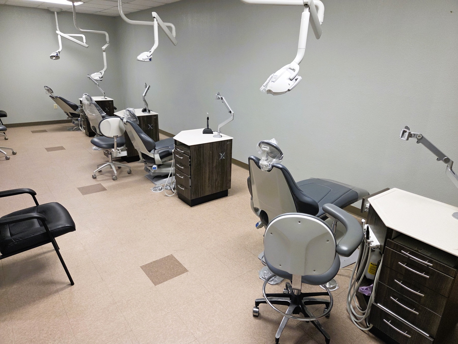 Light grey walls with dark baseboards and tan tiled flooring. Light grey dental procedural chairs anchor the room with an overhead dental procedural light.