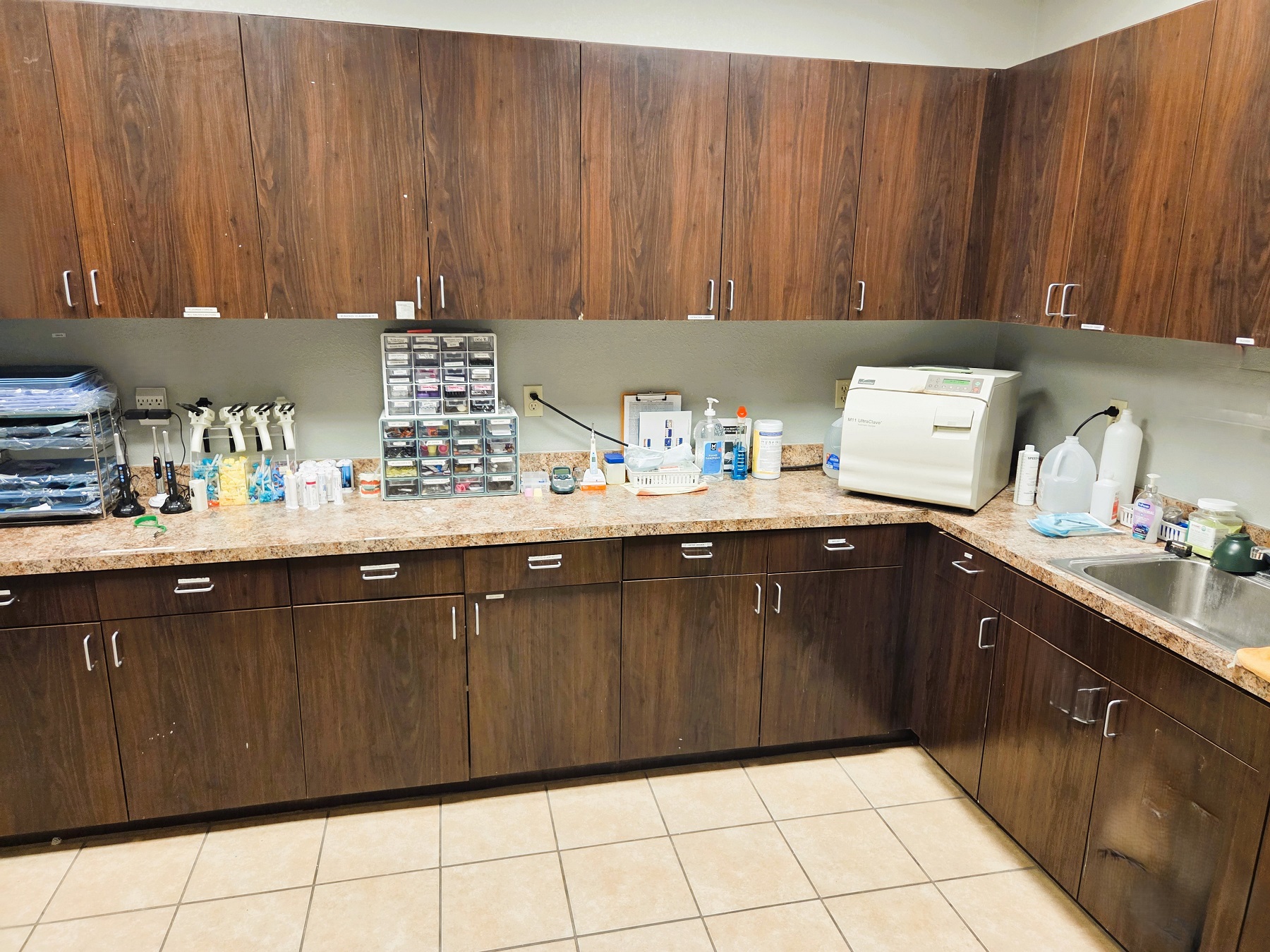 Light grey walls, tan tiled floor, dark stained wood cabinetry with light tan countertops. Lots of storage space for dental lab equipment, tools, and supplies.