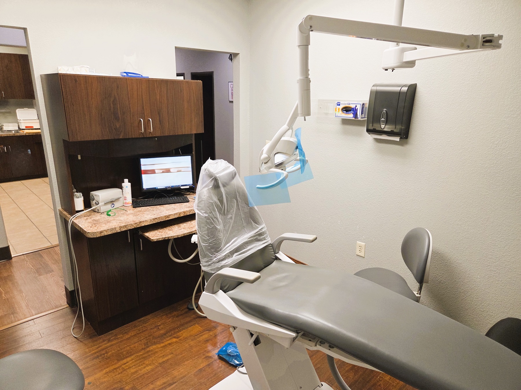 Light grey walls with dark baseboards and dark stained wood-look vinyl flooring. Light grey dental procedural chair anchors the room with an overhead dental procedural light. A dark stained wood cabinet witha light counter top houses a computer and lots of space for storage of dental tools and supplies. Doorway out to the rest of the practice.