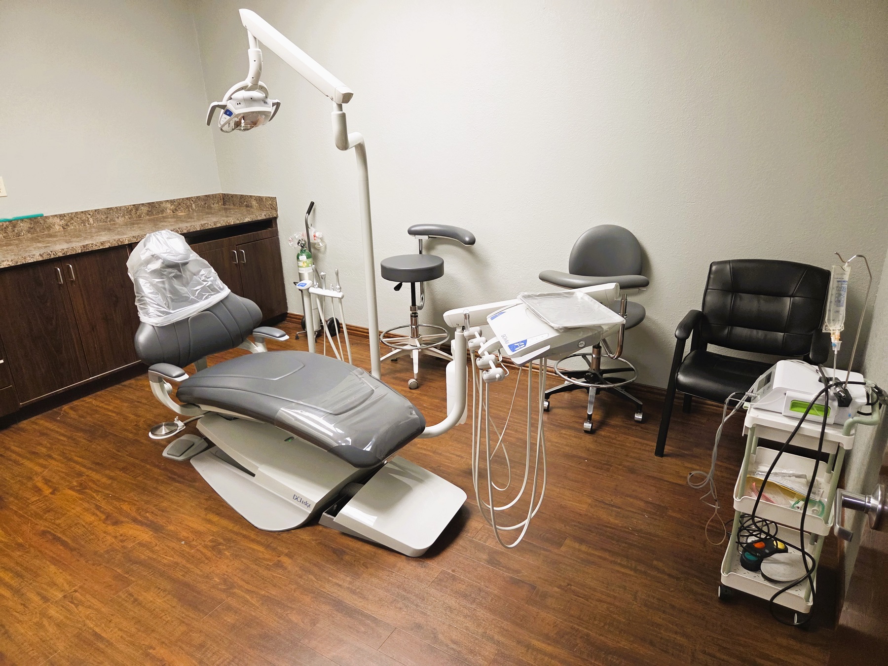 Light grey walls with dark baseboards and dark stained wood-look vinyl flooring. Light grey dental procedural chair anchors the room with an overhead dental procedural light. A dark stained wood cabinet witha light counter top houses a computer and lots of space for storage of dental tools and supplies. Doorway out to the rest of the practice.