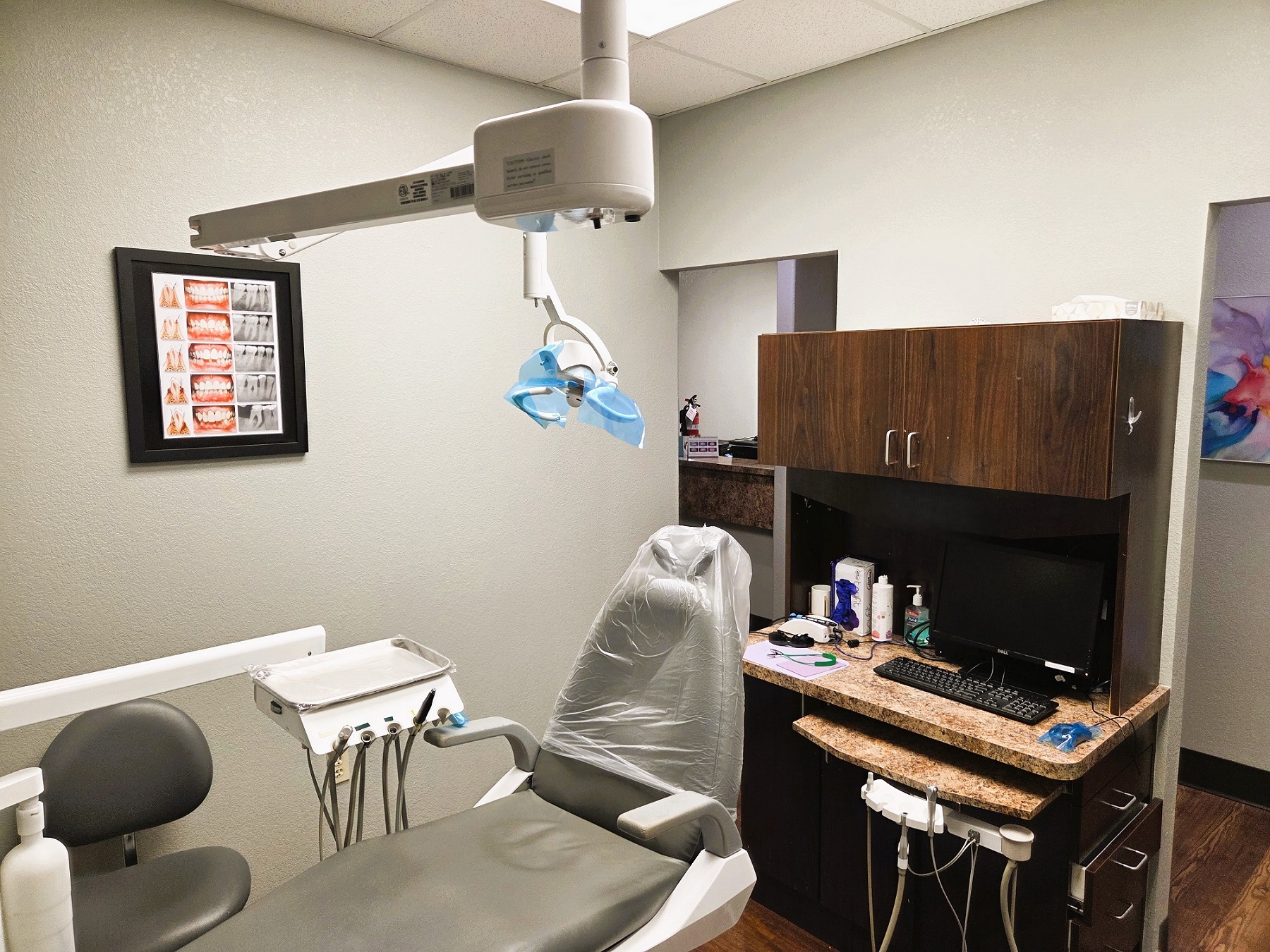 Light grey walls with dark baseboards and dark stained wood-look vinyl flooring. Light grey dental procedural chair anchors the room with an overhead dental procedural light. A dark stained wood cabinet witha light counter top houses a computer and lots of space for storage of dental tools and supplies. Doorway out to the rest of the practice.