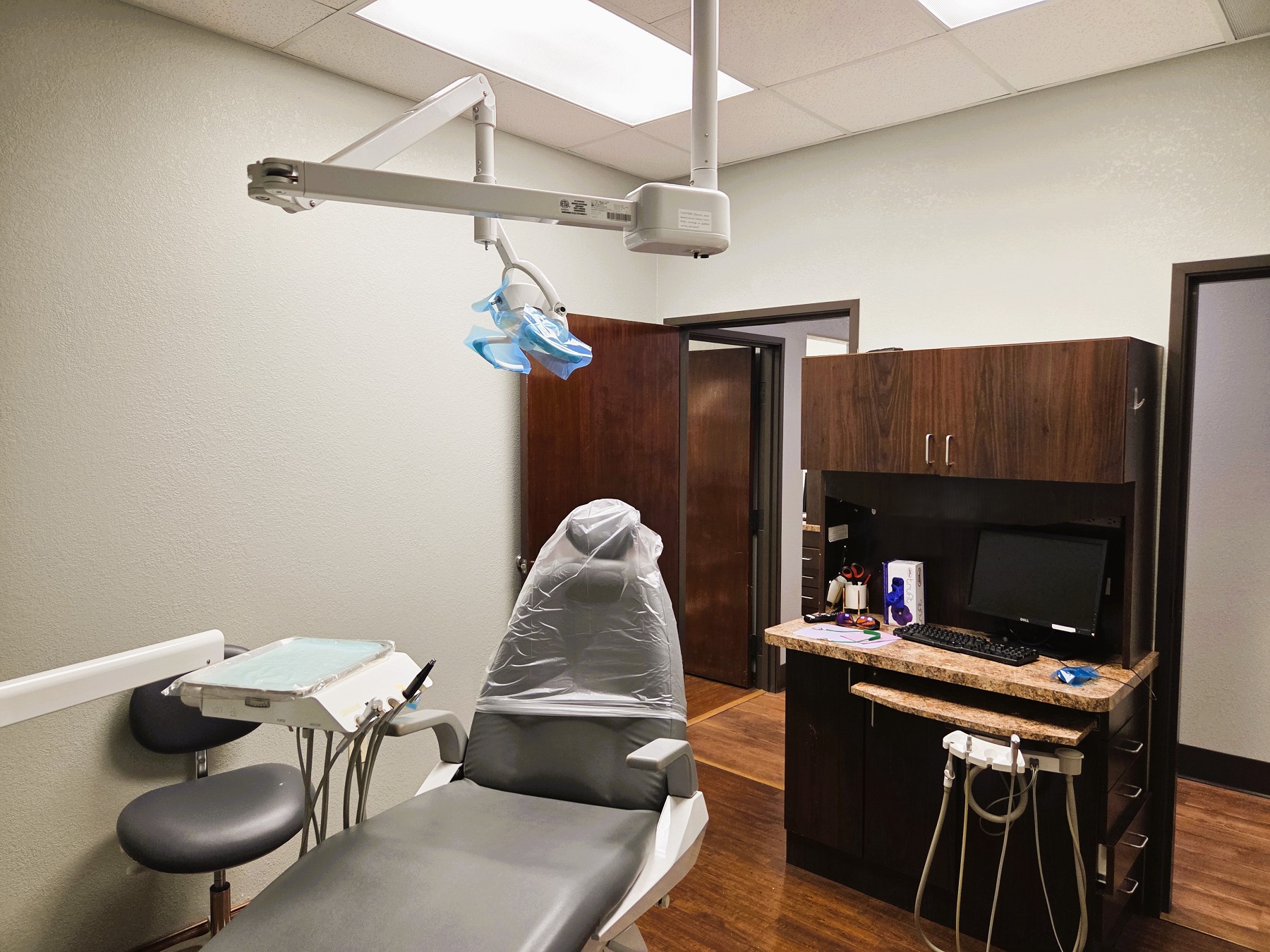 Light grey walls with dark baseboards and dark stained wood-look vinyl flooring. Light grey dental procedural chair anchors the room with an overhead dental procedural light. A dark stained wood cabinet witha light counter top houses a computer and lots of space for storage of dental tools and supplies. Doorway out to the rest of the practice.