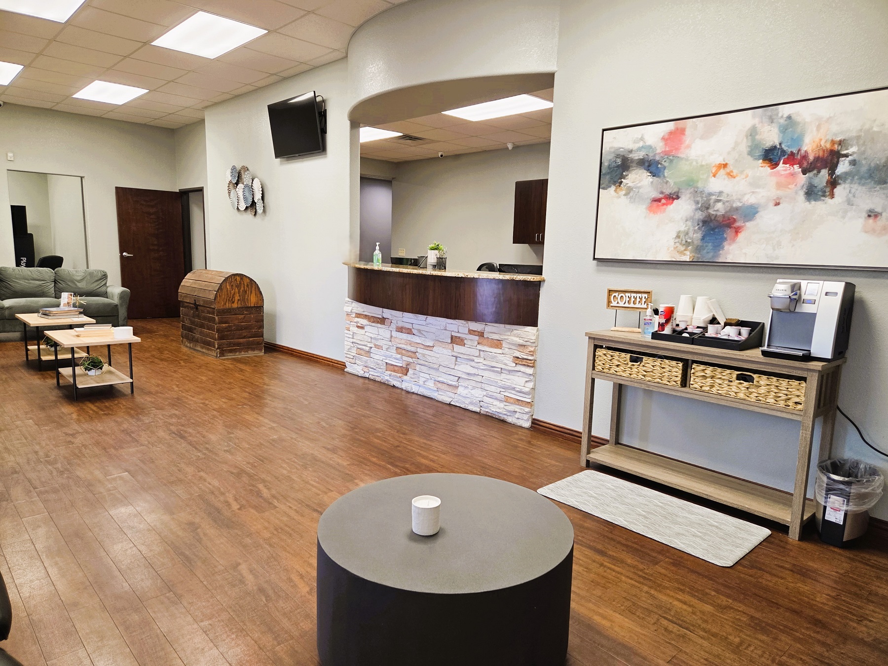 Light grey walls with dark baseboards and dark, stained, wood-look flooring. Built in arched reception window with stone veneer below the desk and looking into the front office. Large artwork on the walls, coffee and end tables and a coffee station. Over stuffed seats and a doorway to the back of the practice.