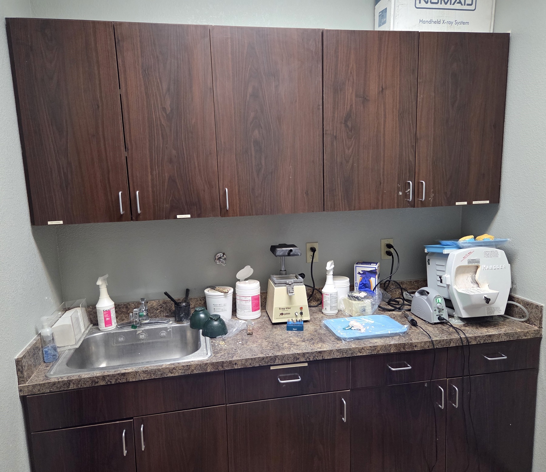 Light grey walls, tan tiled floor, dark stained wood cabinetry with light tan countertops. Lots of storage space for dental sterilization equipment, tools, and supplies.
