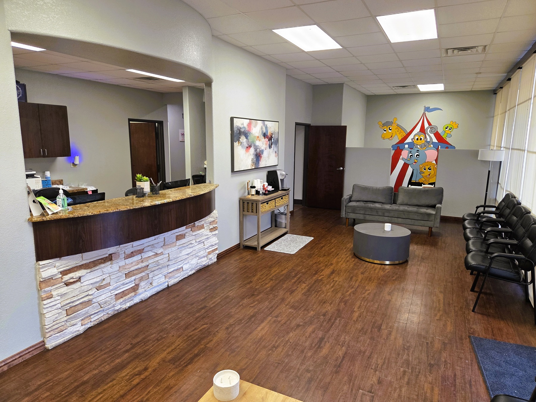 Light grey walls with dark baseboards and dark, stained, wood-look flooring. Built in arched reception window with stone veneer below the desk and looking into the front office. Large artwork on the walls, coffee and end tables and a coffee station. Over stuffed seats and a doorway to the back of the practice.