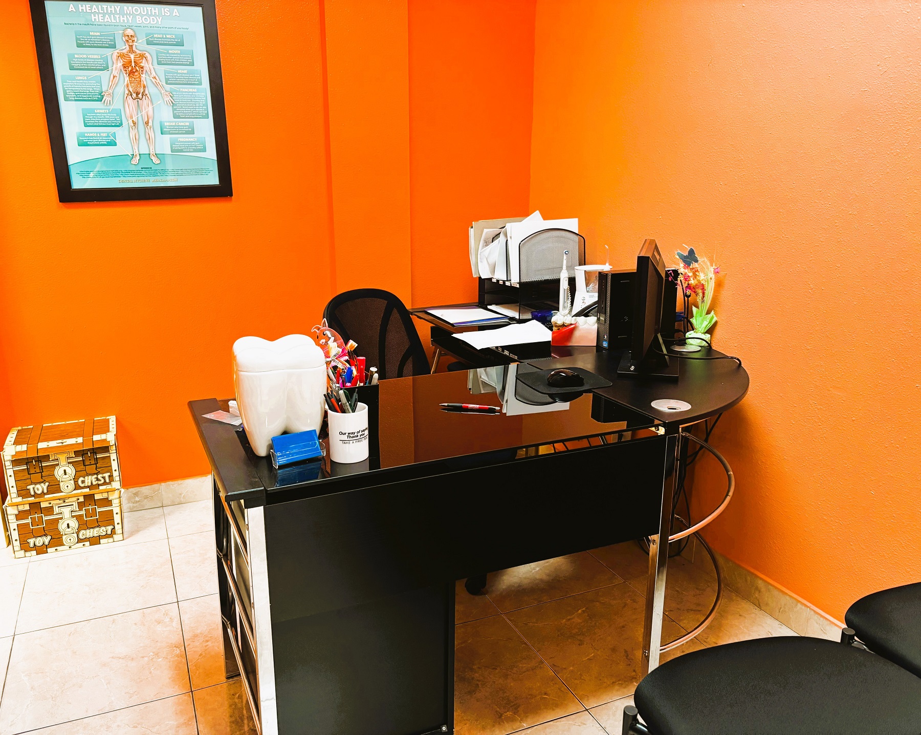 Bright and light colored walls with white trim and large grey tile flooring. An "L" shaped black desk holds office equipment and supplies with a black office chair pulled up to it.