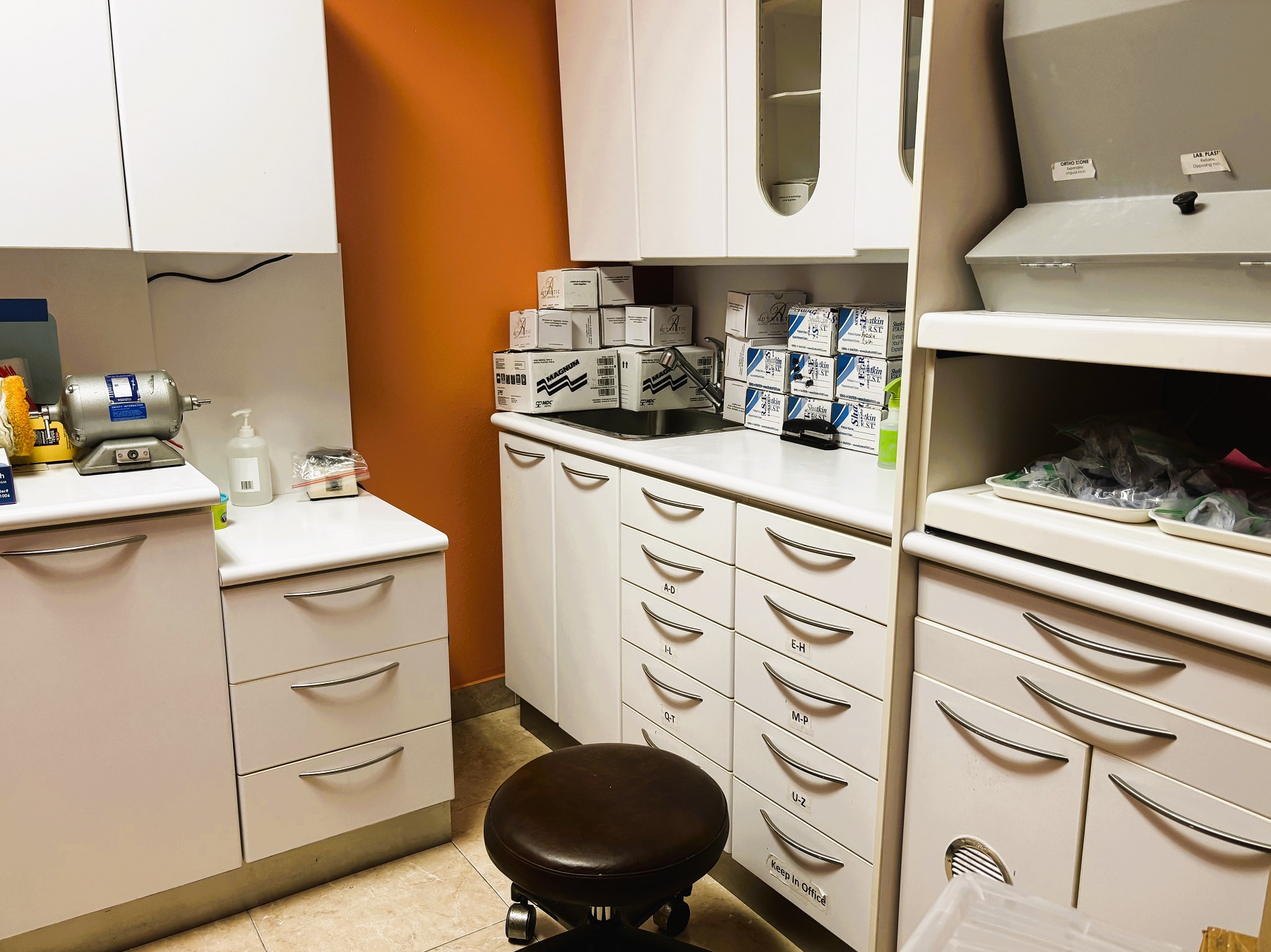 Bright and light colored walls with large grey tile flooring and grey cabinetry with matching counter tops. Lots of dental lab equipment and supplies.