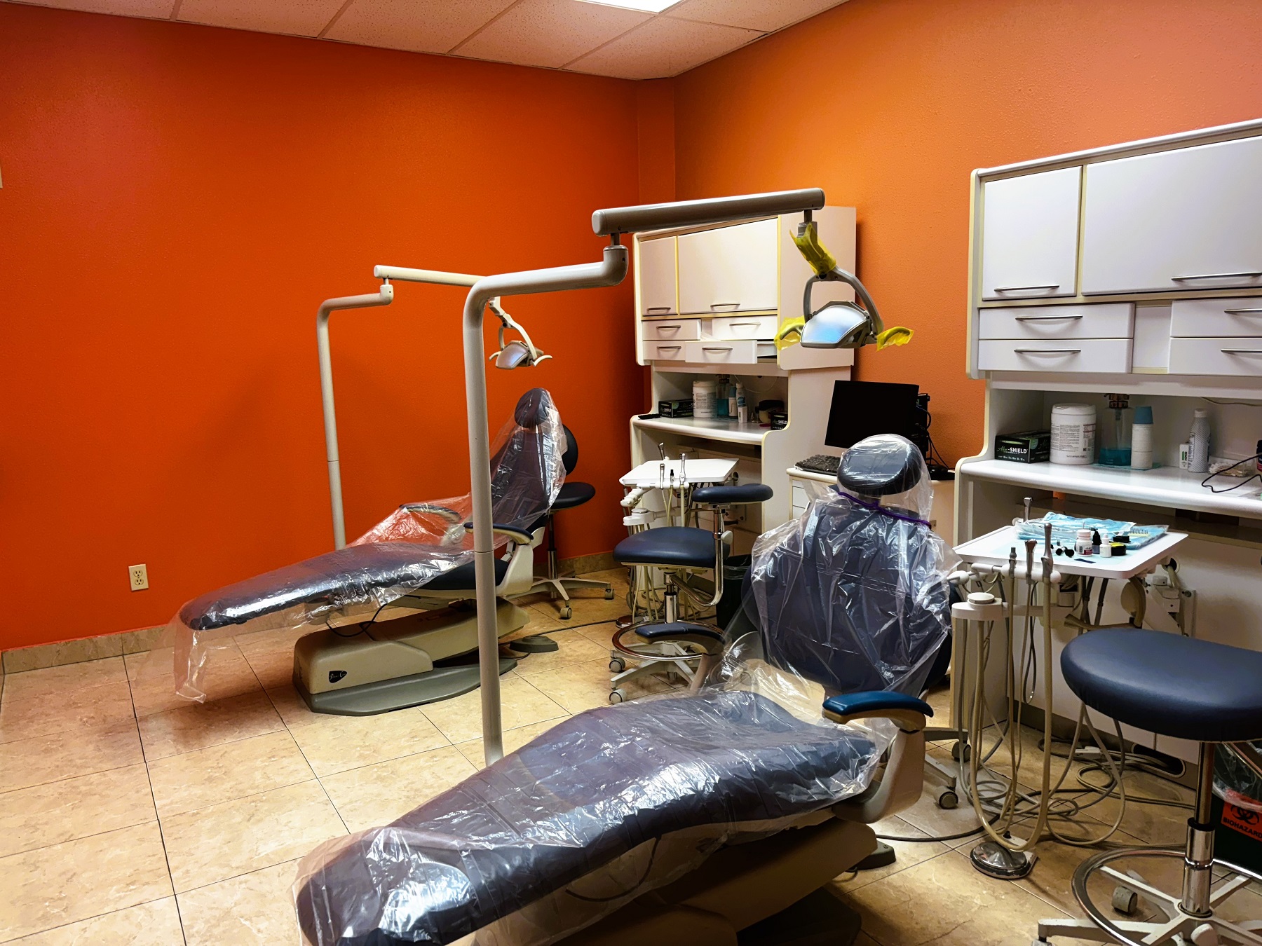 bright and light colored walls with white trim and large grey tile flooring. Dental procedural chair anchors the room. Light cabinetry sits against a wall to hold all the dental tools, supplies, and equipment. Overhead dental procedural lighting sits next to the dental chair.