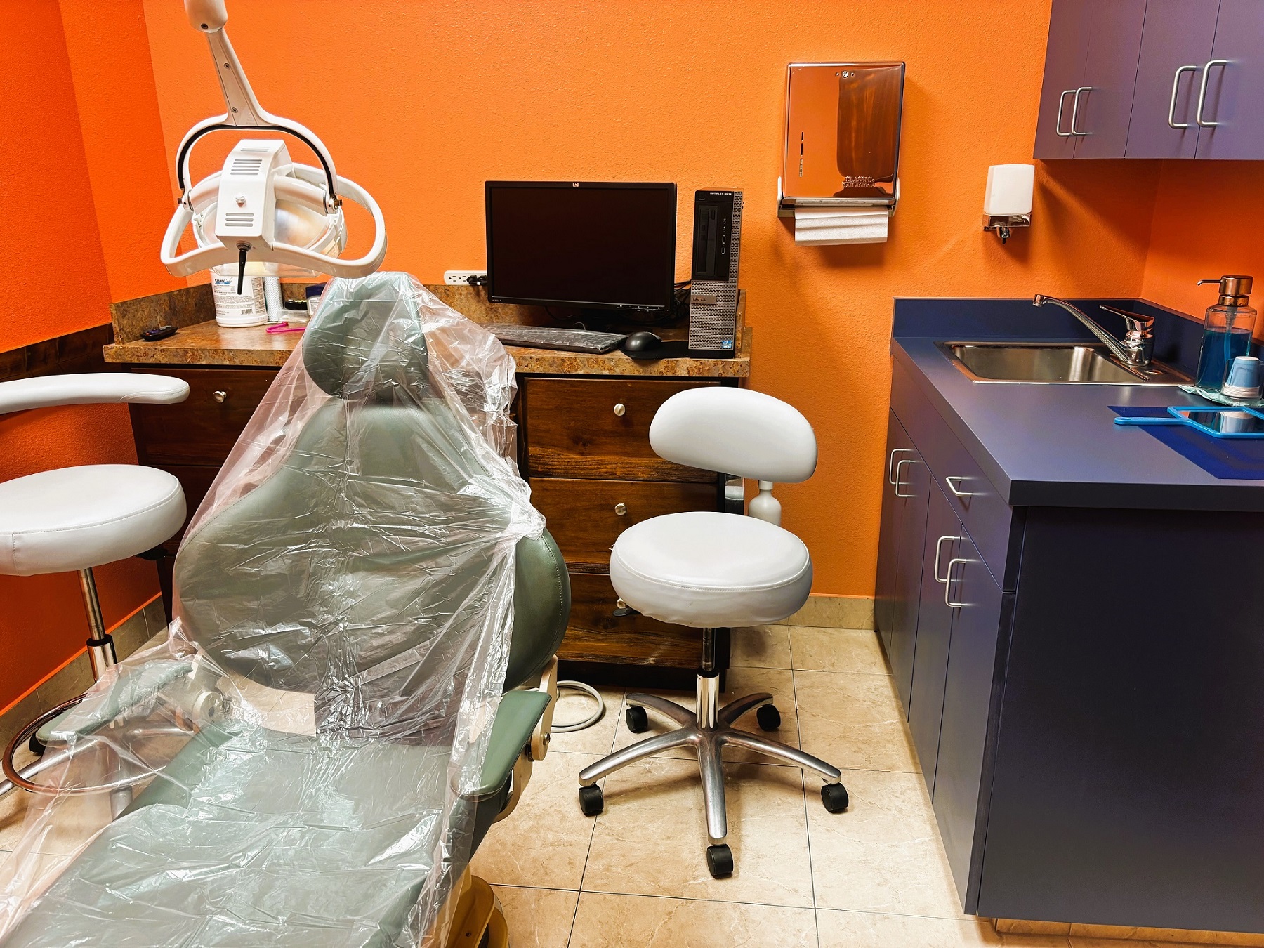 bright and light colored walls with white trim and large grey tile flooring. Dental procedural chair anchors the room. Light cabinetry sits against a wall to hold all the dental tools, supplies, and equipment. Overhead dental procedural lighting sits next to the dental chair.