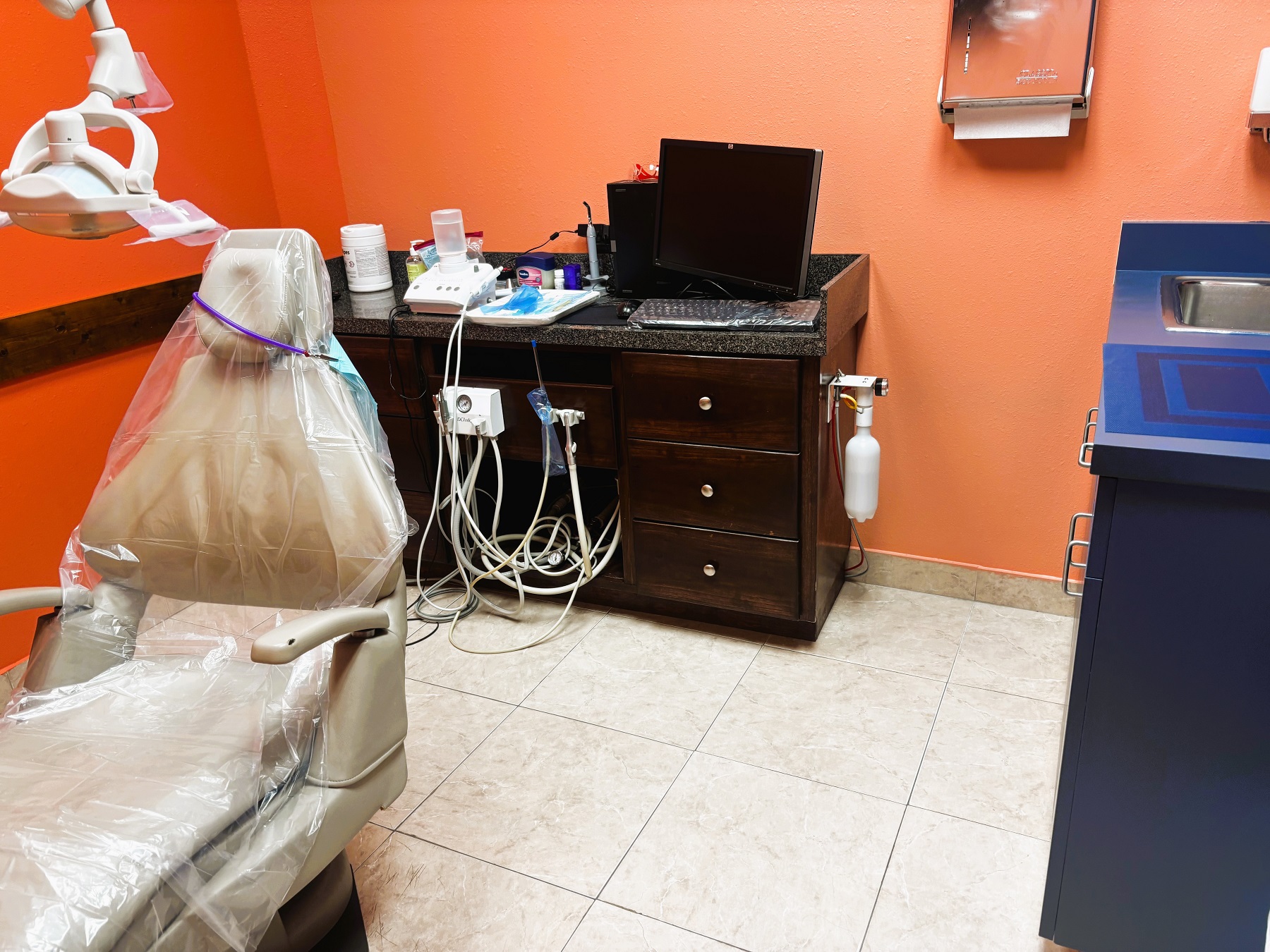 bright and light colored walls with white trim and large grey tile flooring. Dental procedural chair anchors the room. Light cabinetry sits against a wall to hold all the dental tools, supplies, and equipment. Overhead dental procedural lighting sits next to the dental chair.
