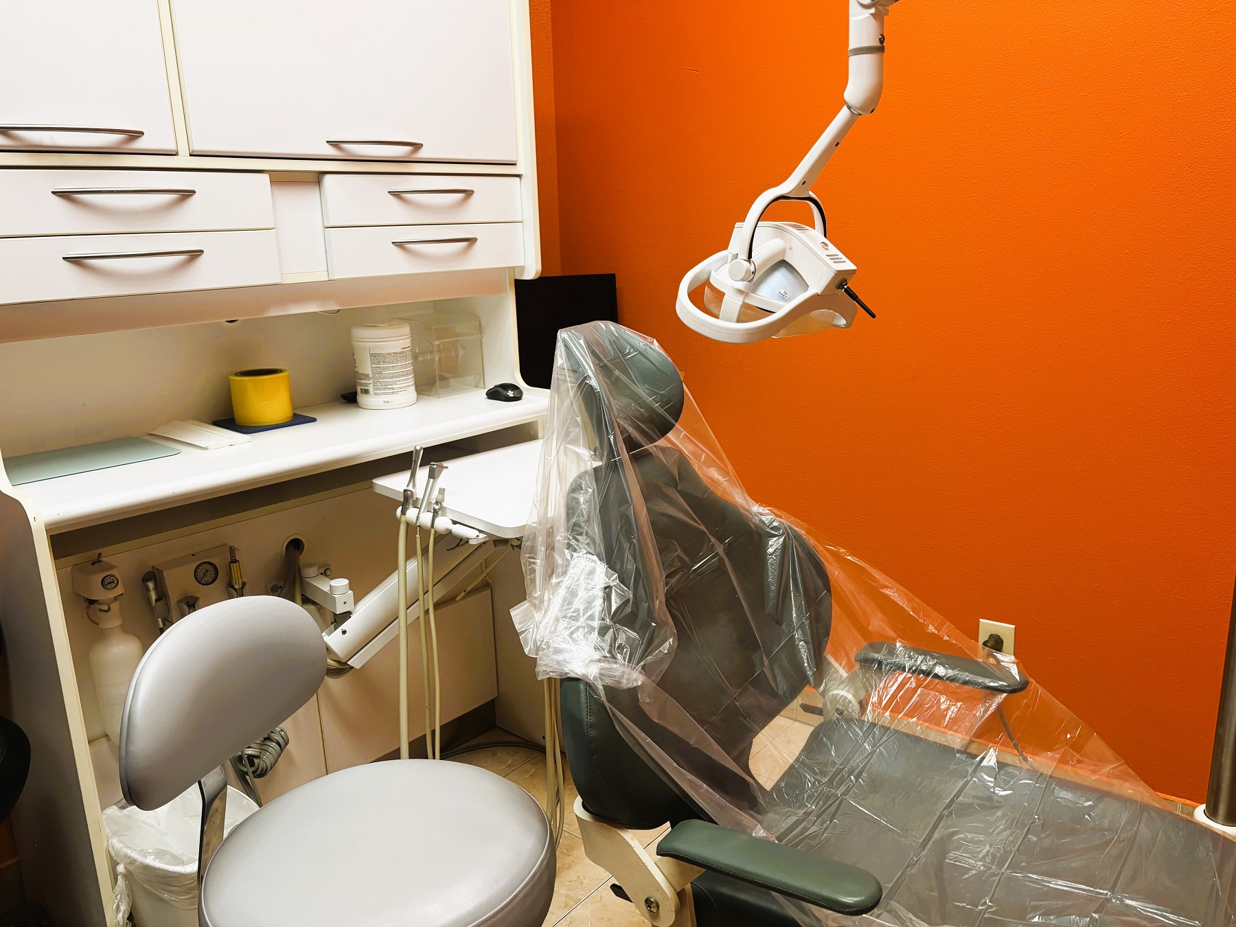 bright and light colored walls with white trim and large grey tile flooring. Dental procedural chair anchors the room. Light cabinetry sits against a wall to hold all the dental tools, supplies, and equipment. Overhead dental procedural lighting sits next to the dental chair.