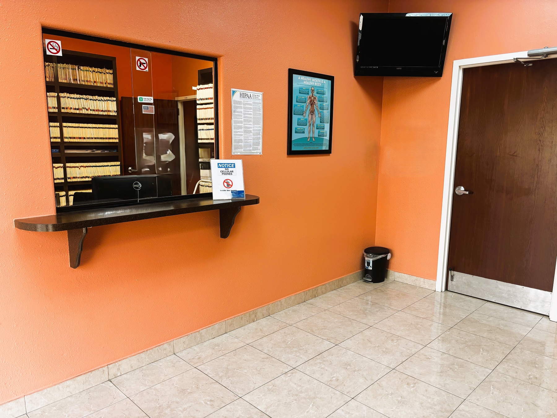 Bright and light walls, white trim, and large grey tile flooring. Dark stained wood door leading to the back of the practice. Large flat screen tv is mounted up in the corner of the room and a large reception window with tinted glass and a black rounded counter.