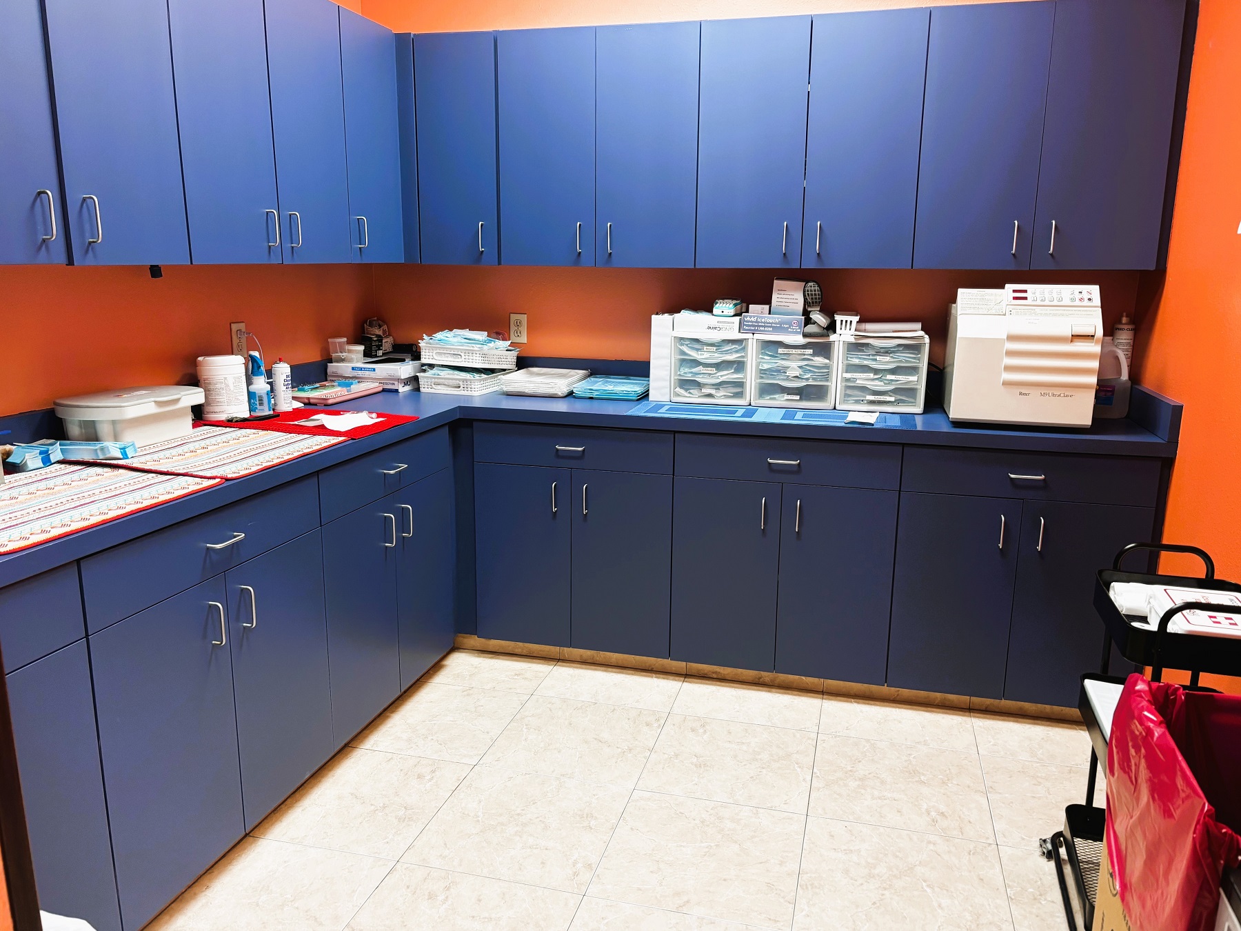 Bright and light colored walls with large grey tile flooring. Blue cabinets with matching counter tops hold lots of dental sterilization equipment and supplies.