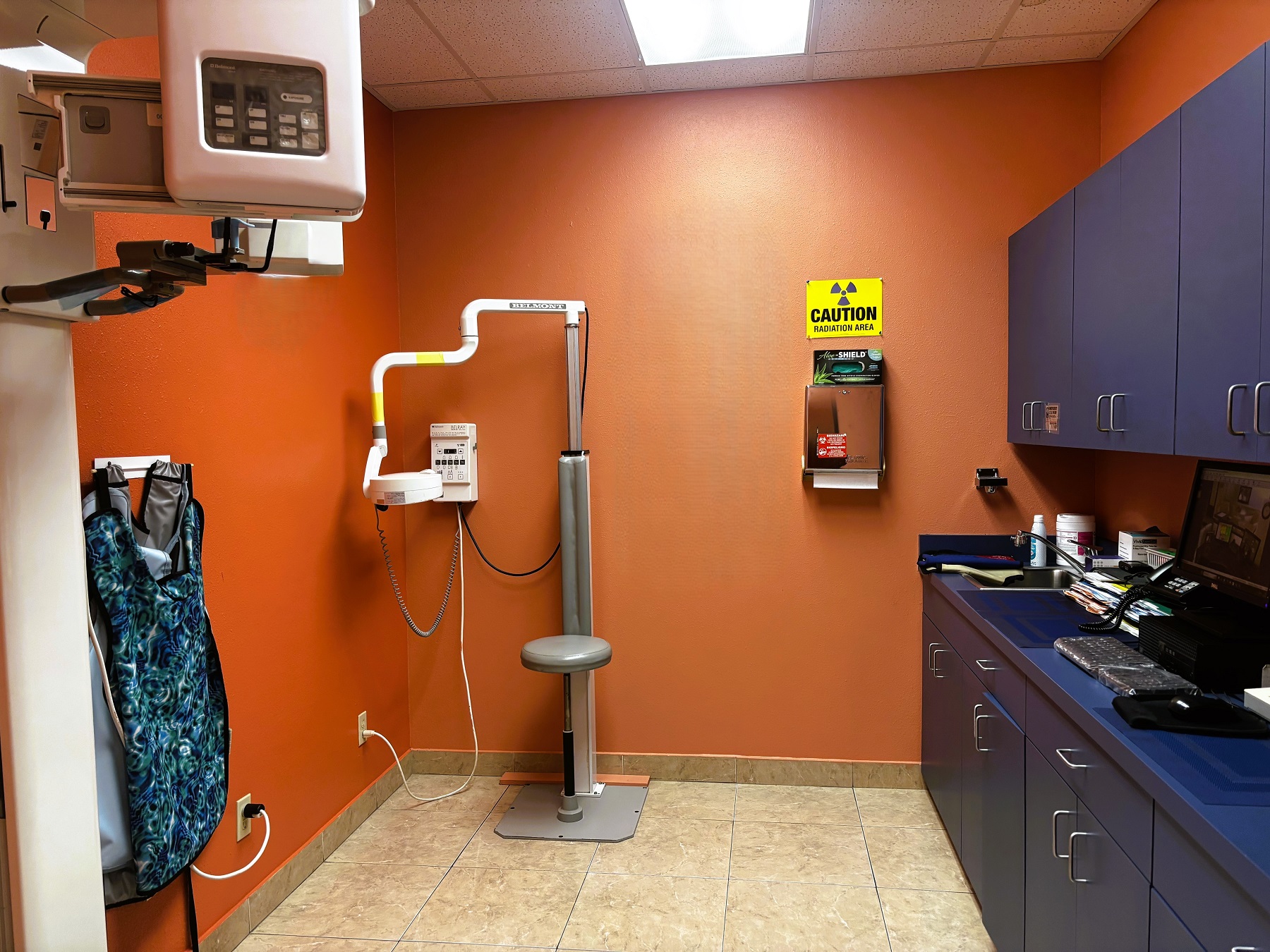 Bright and light colored walls with large grey tile flooring. Dental x-ray equipment sits against the wall.