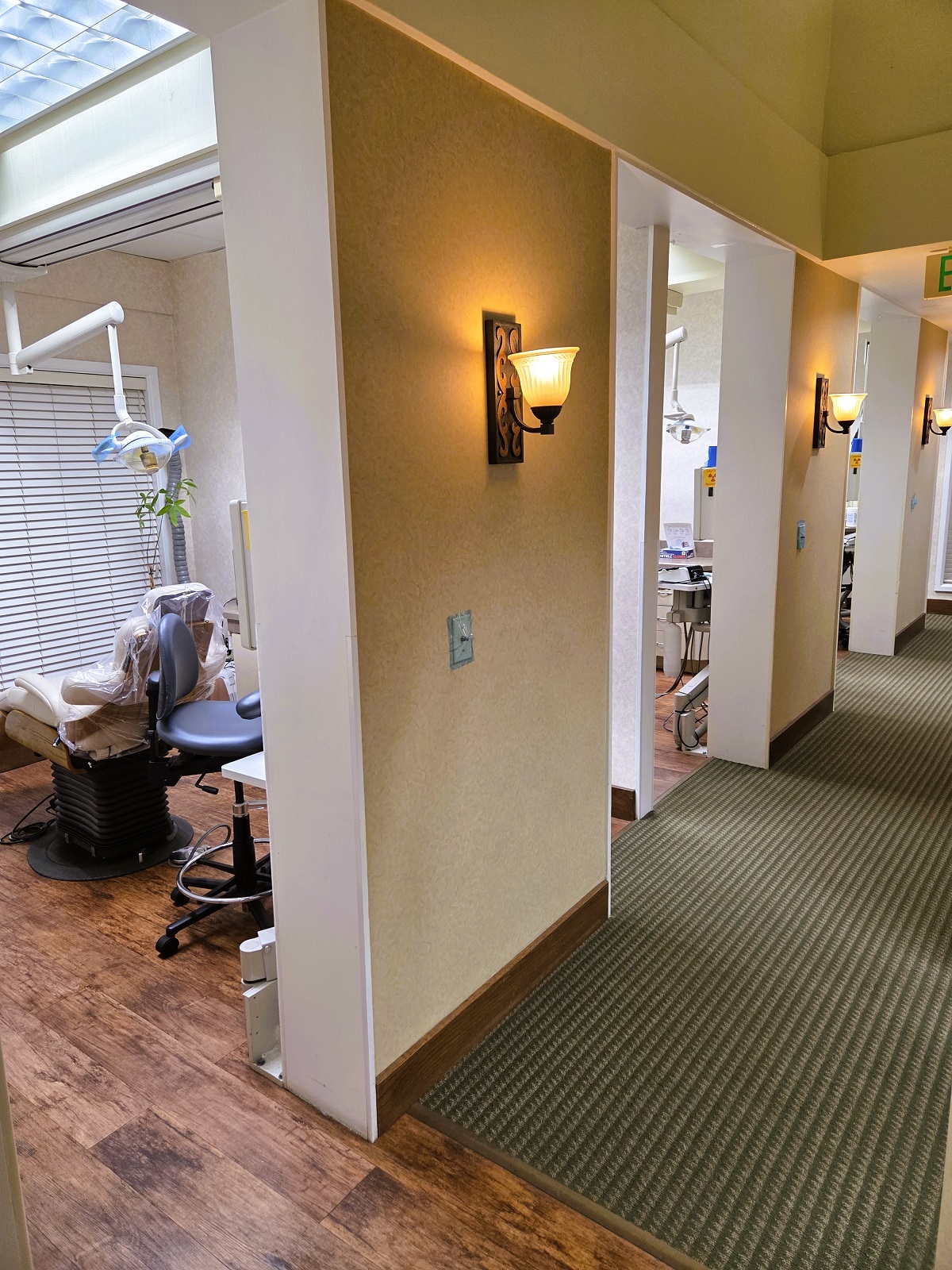 Tan walls with dark stained wood extra wide baseboards and light sconces leading down the hallway. Doorways open to the different operatories and rooms of the practice. Dark wood flooring and similar tones of short Berber carpet go into the operatories and run down the hallway.