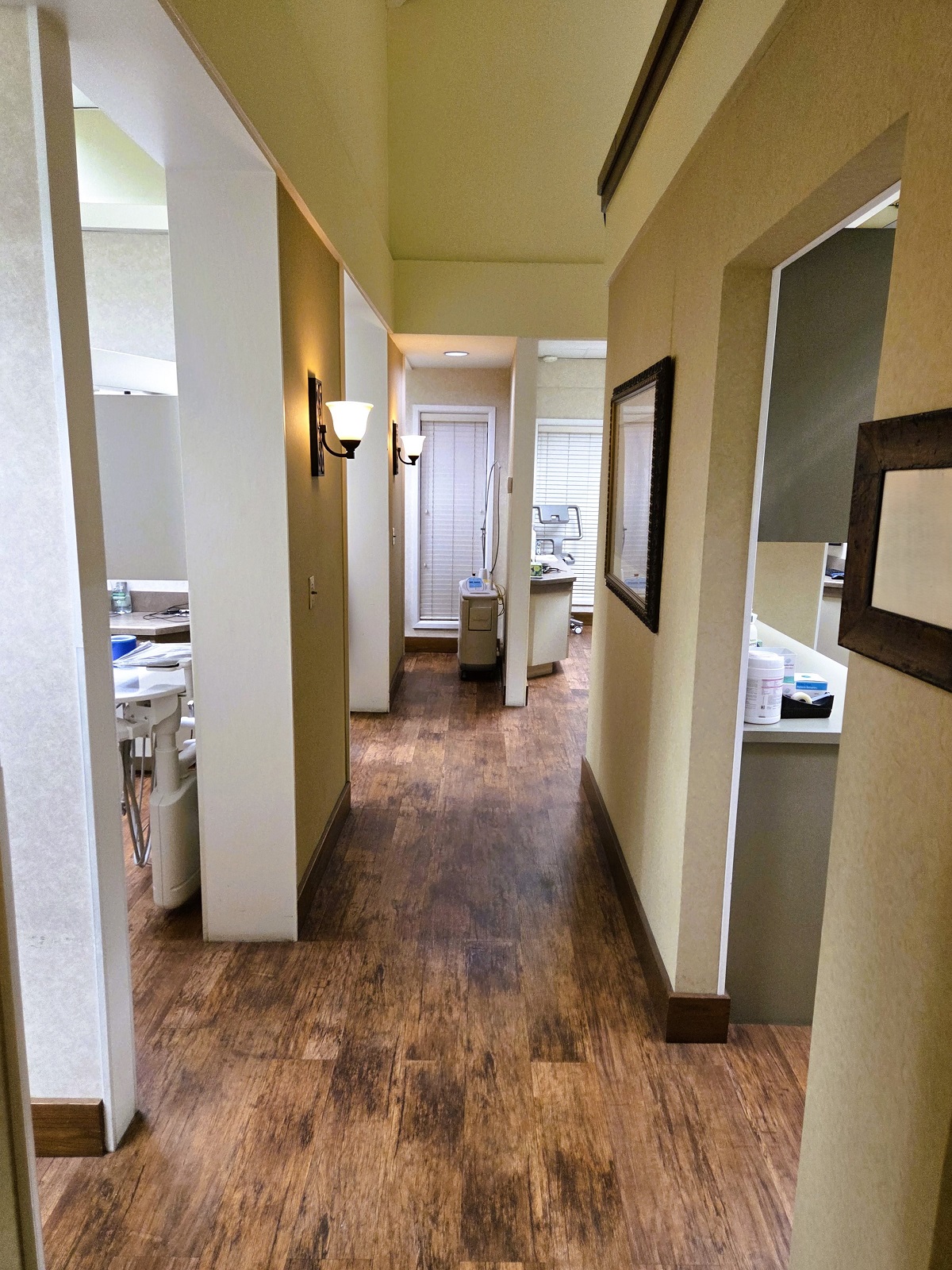 Tan walls with dark stained wood extra wide baseboards and light sconces leading down the hallway. Doorways open to the different operatories and rooms of the practice. Dark wood flooring and similar tones of short Berber carpet go into the operatories and run down the hallway.