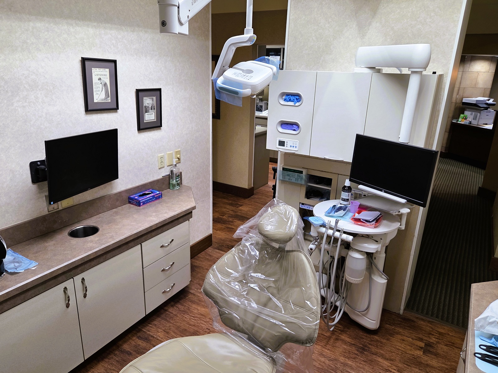 Light tan walls with matching color cabinetry and slightly darker countertops. Dark stained wood baseboards and trim. Dark stained wood flooring. A tan dental chair anchors the room with an overhead dental procedural light. Portable trays hold lots of lots of dental tools, supplies, and equipment.