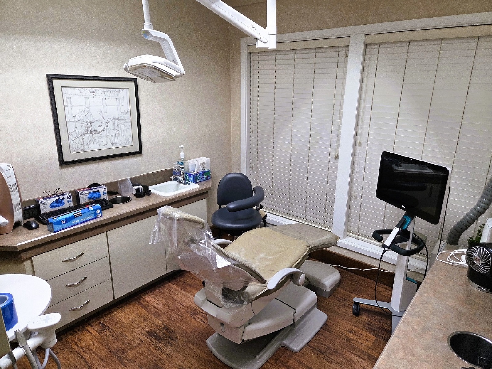Light tan walls with matching color cabinetry and slightly darker countertops. Dark stained wood baseboards and trim. Dark stained wood flooring. A tan dental chair anchors the room with an overhead dental procedural light. Portable trays hold lots of lots of dental tools, supplies, and equipment.
