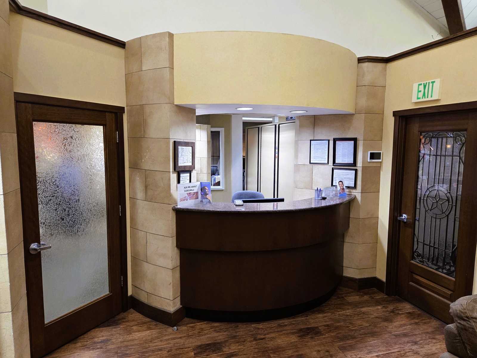 Two tone curved wall with large tiles, wall paper and paint all in neutral tan shades with dark stained wood. A matching dark stained wood reception desk is built into the curved wall with overhead can lighting shining down on the matching dark counter top. This looks back into the front office of the practice. To the left a large dark wood door with obscure glass leads to the back of the practice.