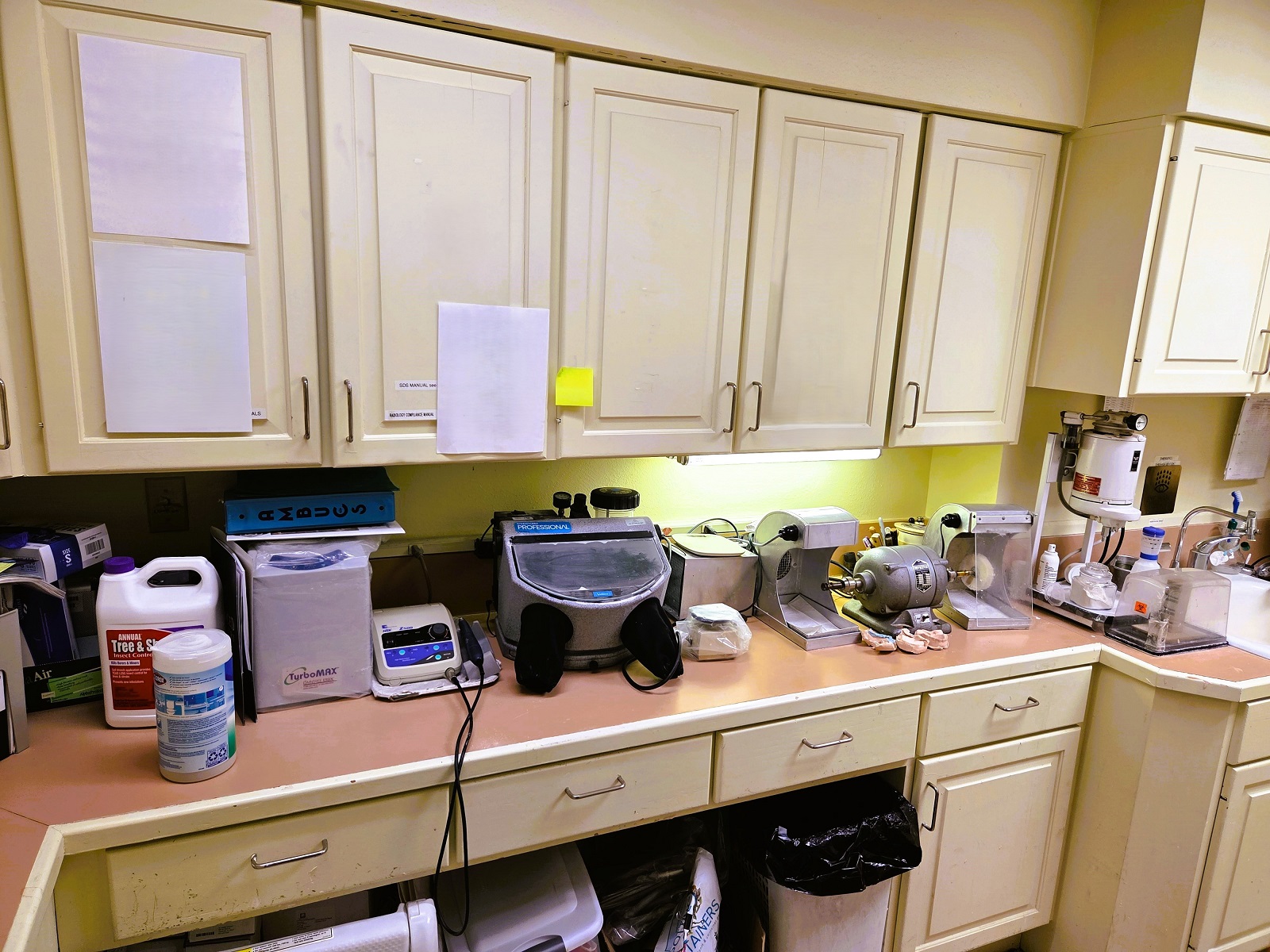 Light tan walls with matching color cabinetry and countertops. Dark wood look flooring. Upper cabinets and the counter with a double sink hold lots of dental supplies and tools.