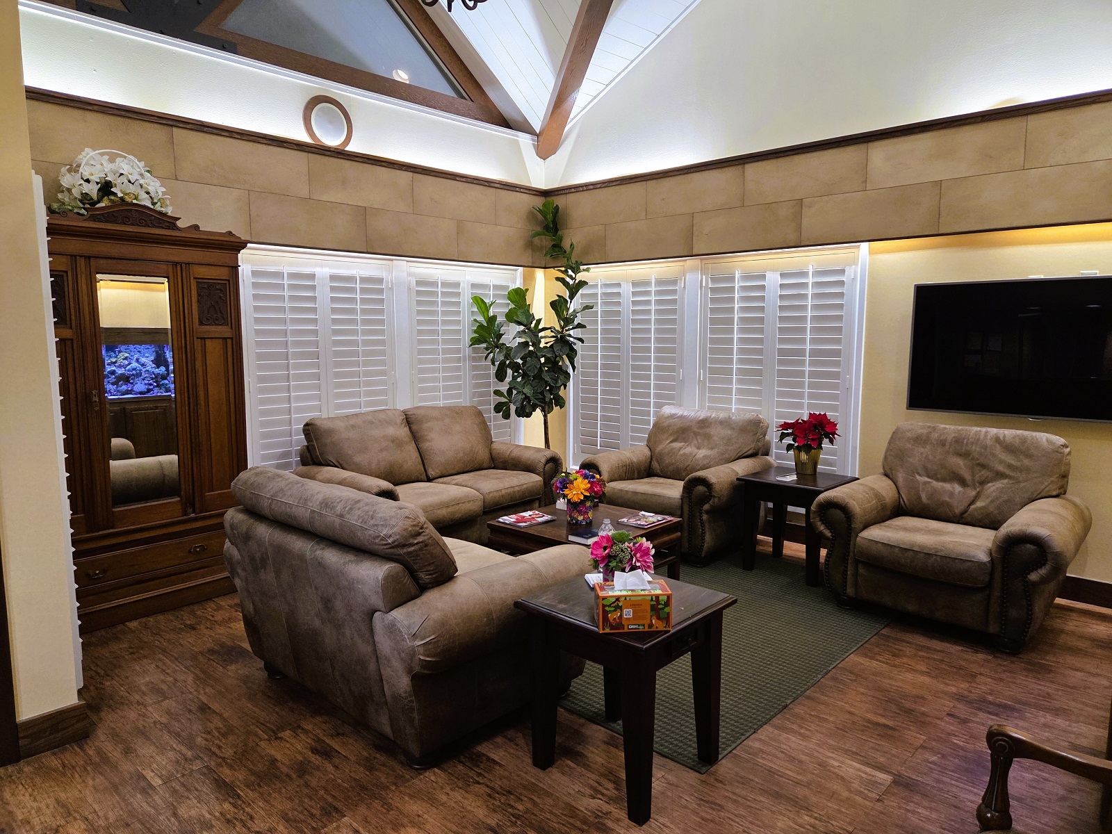 Two tone walls in neutral tans and white with dark stained baseboards and trim work. White plantation shutters cover large windows on the back wall with large over-stuffed leather love seats and chairs surrounding a coffee table and with end tables holding magazines between them sitting on dark stained wood flooring. Vaulted ceilings with huge windows and beams give the room a rich and luxurious look.