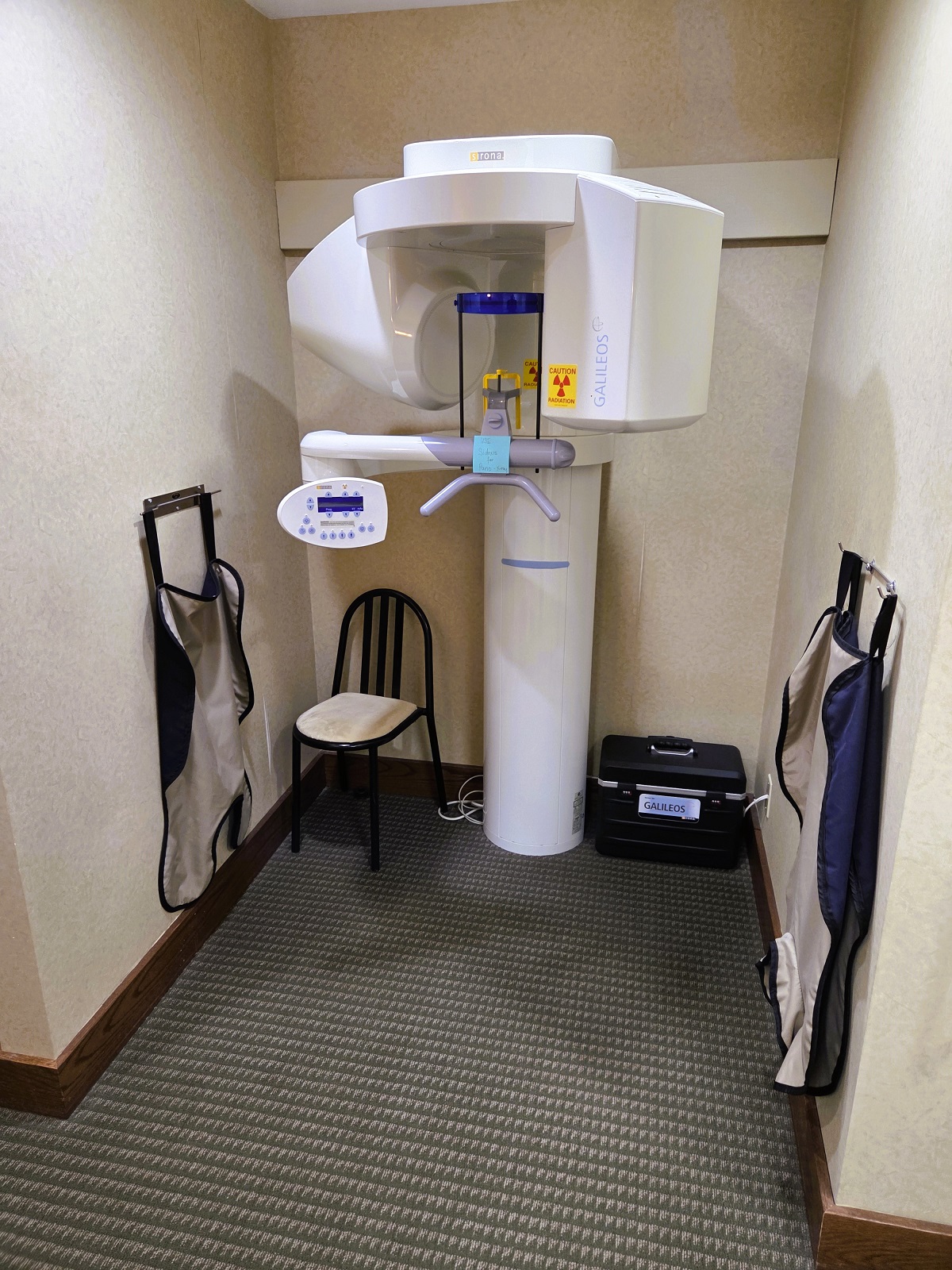 Light tan walls with dark stained baseboards and multi toned brown Berber carpet. A large dental x-ray machine sits in the alcove.