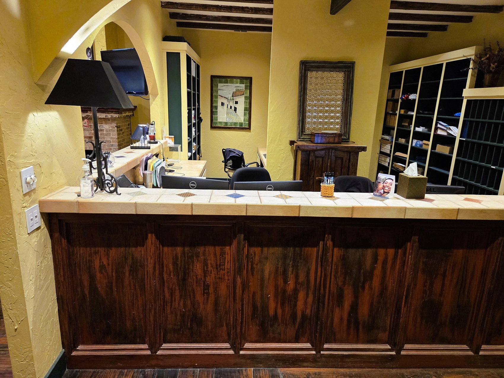 Dark stained wood panels sit under a tan tile countertop looking back into the front office with light colored walls with arches over the countertop. Builtin matching wood bookcases hold office equipment. Office equipment and supplies sit on the desktop that's built into the front office.