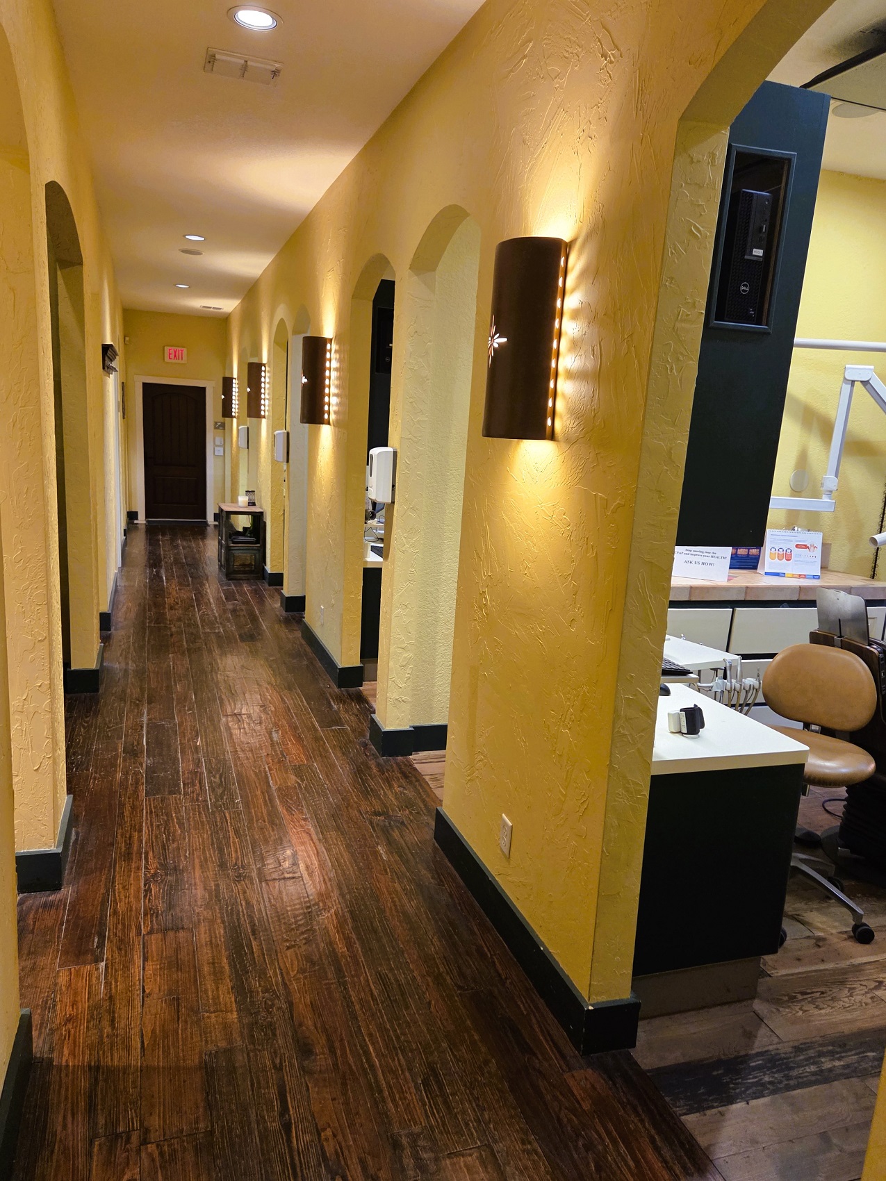 Hand scraped, dark stained wood flooring stretches the length of the hallway. Light colored walls with dark light sconces framing doorways all the way down the hallway. Can lights in the ceiling bring additional lighting to the situation.