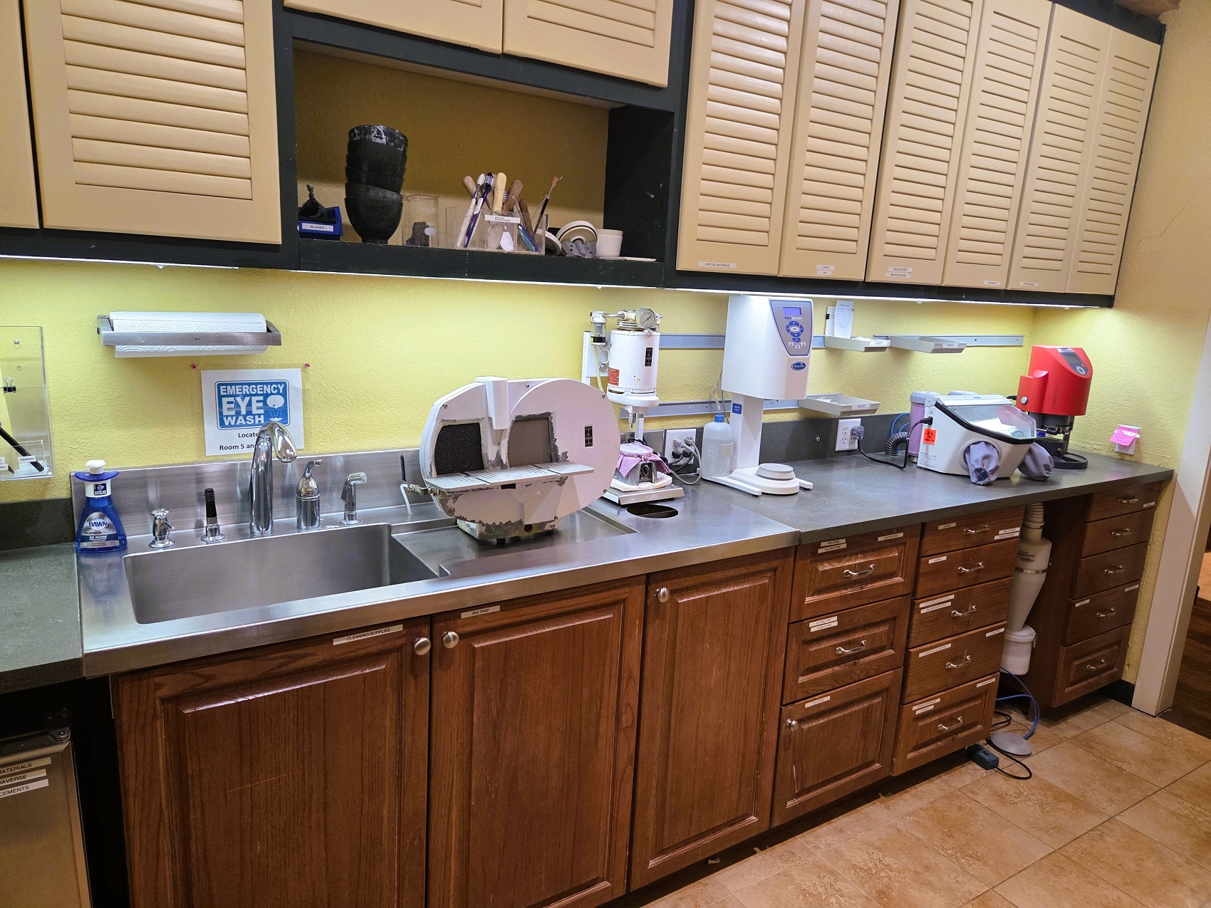 Light colored walls, dark stained wood trim, and large terracotta colored tile flooring. Dark stained cabinetry with black countertop and stainless steal sink. Upper cabinets with matching dark stained cabinet bodies have lighter colored louvered doors. These hold lots of dental equipment, tools and supplies.