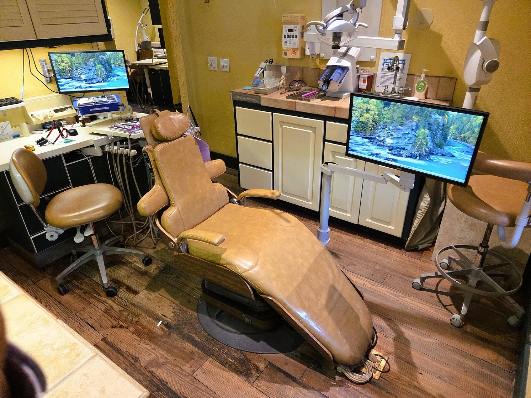 Dark stained, hand scraped wood flooring with light colored walls. A cushioned dental procedural chair anchors the room with overhead dental lighting. Dark framed cabinetry with light colored doors and drawer fronts holds lots of dental equipment and supplies.