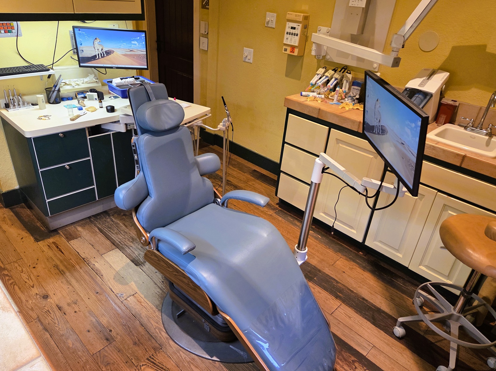 Dark stained, hand scraped wood flooring with light colored walls. A cushioned dental procedural chair anchors the room with overhead dental lighting. Dark framed cabinetry with light colored doors and drawer fronts holds lots of dental equipment and supplies.