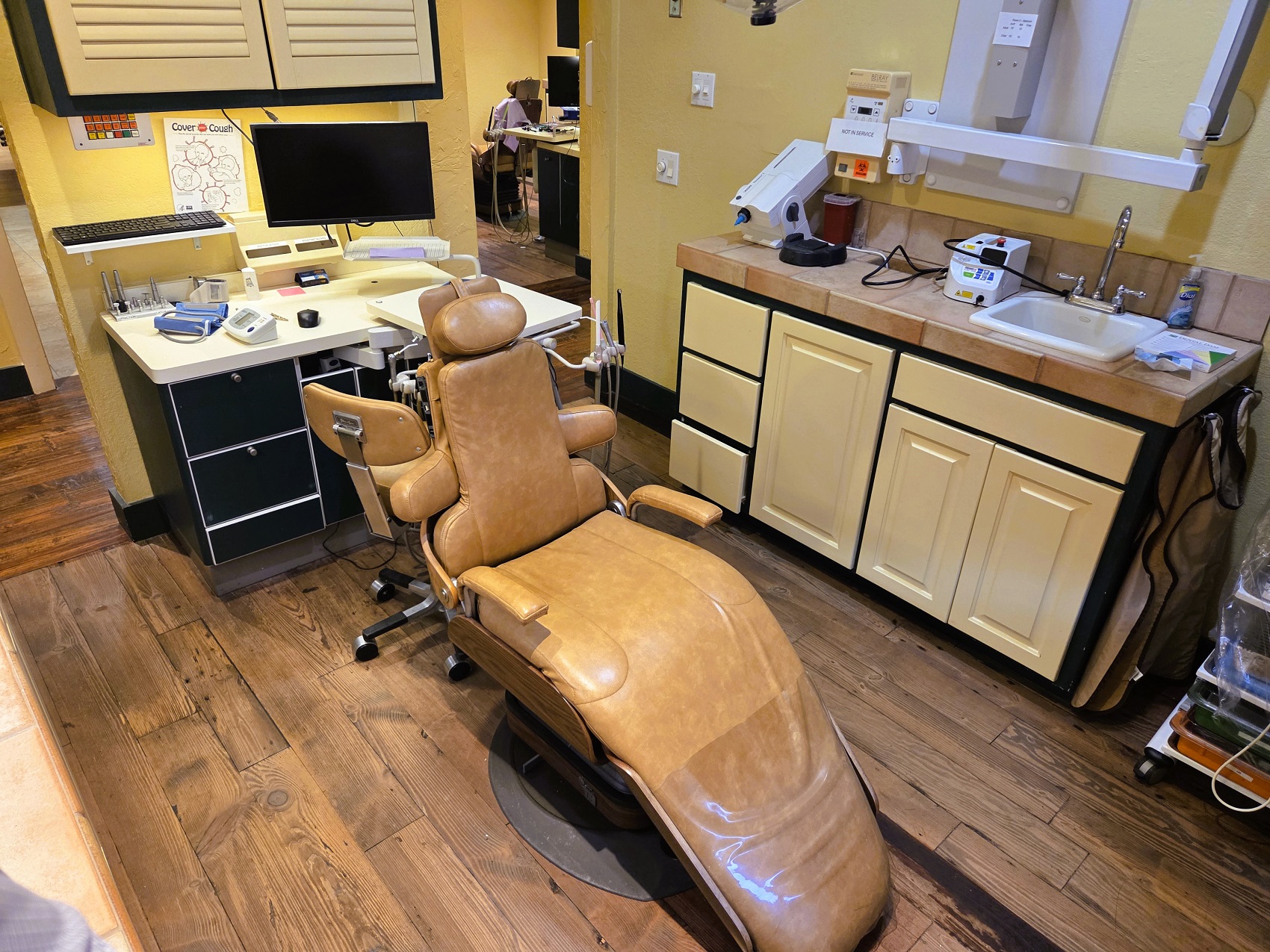 Dark stained, hand scraped wood flooring with light colored walls. A cushioned dental procedural chair anchors the room with overhead dental lighting. Dark framed cabinetry with light colored doors and drawer fronts holds lots of dental equipment and supplies.