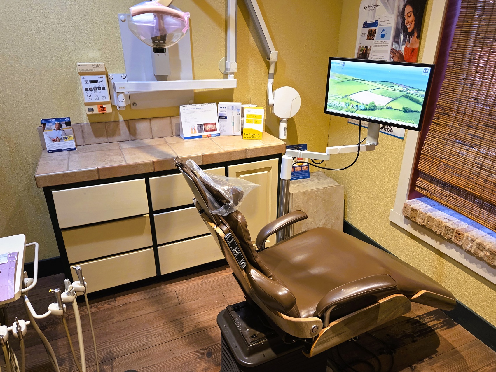 Dark stained, hand scraped wood flooring with light colored walls. A cushioned dental procedural chair anchors the room with overhead dental lighting. Dark framed cabinetry with light colored doors and drawer fronts holds lots of dental equipment and supplies.