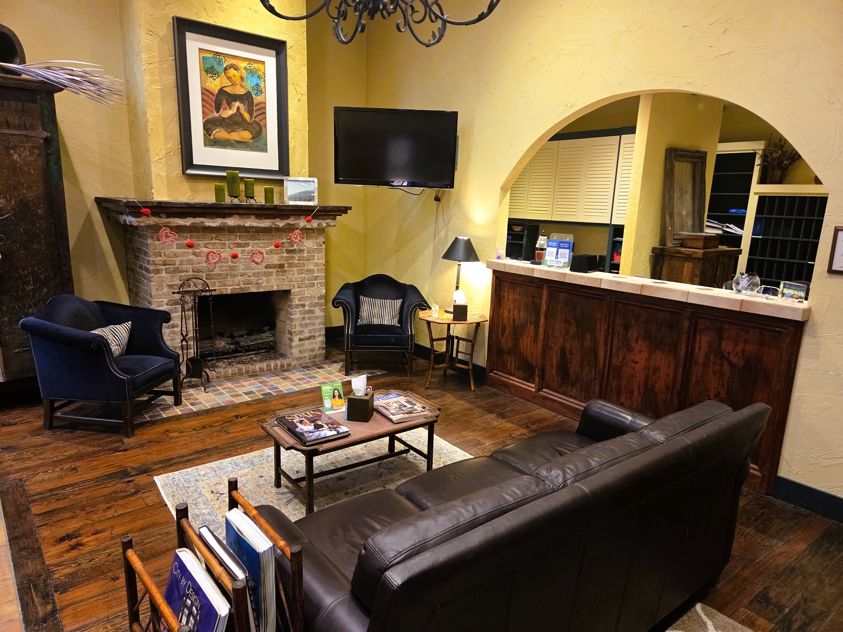 Light colored walls with dark stained wood trim and dark stained hand scraped hardwood flooring. A brick fireplace with a dark stained wood mantel has a large framed piece of art hanging over it. A large, flat screen TV is mounted in the corner to the right. Matching wood is on the wall under an arched reception desk with a light tan tile countertop. an overstuffed sofa with two matching arm chairs flanking the fireplace sit around a coffee table that holds magazines. The arm chair closest to the reception counter has a small side table next to it with a reading lamp on it. A large armoire sits behind the other arm chair. Office equipment and furniture can be seen behind the reception counter.