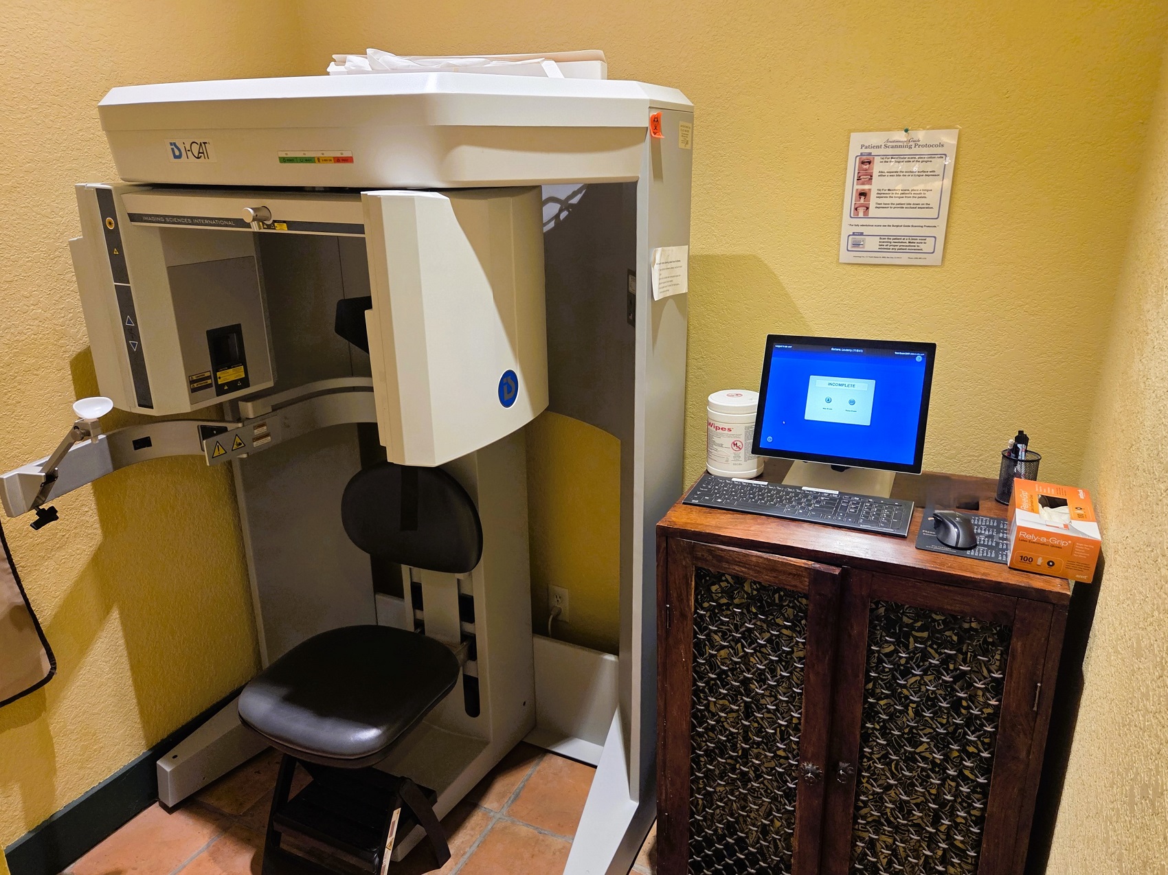 Light colored walls with dark stained wood trim and large terracotta tile flooring> A large dental x-ray machine sits on the left and a dark stained cabinet sits to the right of it holding a computer and dental supplies and equipment.