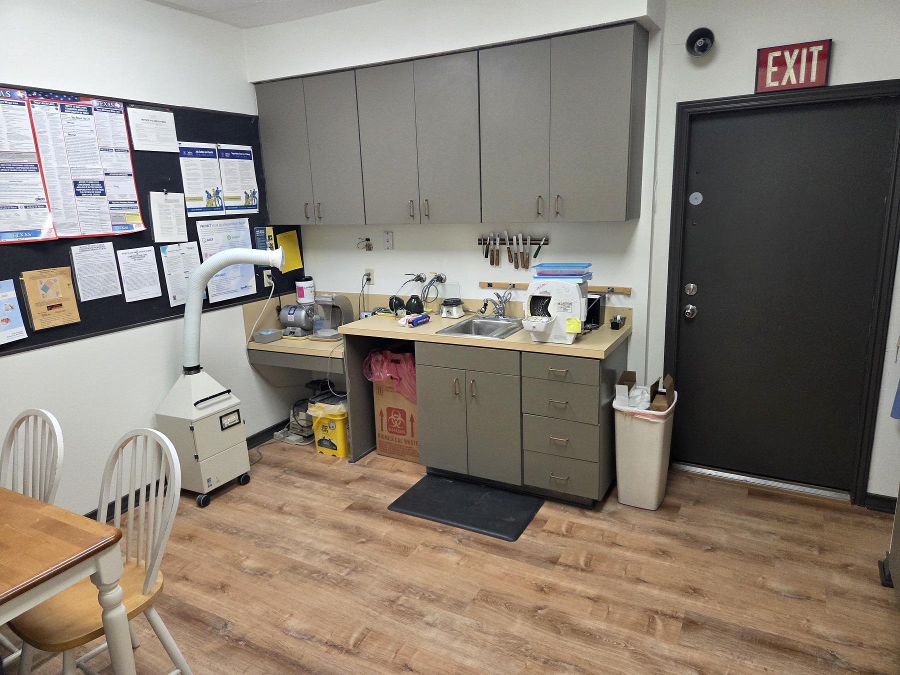 light walls with dark stained baseboards and medium stained wood flooring. A wood table with light wood top and white legs and matching chairs. Dark cabinets with an orangy counter top holds lots of equipment and supplies.