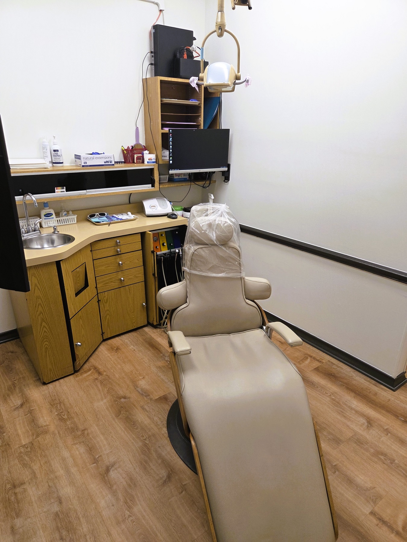 Light walls with dark stained baseboards and chair rail and medium stained wood flooring. A cushioned dental procedural chair anchors the room with an overhead dental light. A cabinet with sink, shelves hold lots of equipment, tools, and supplies.