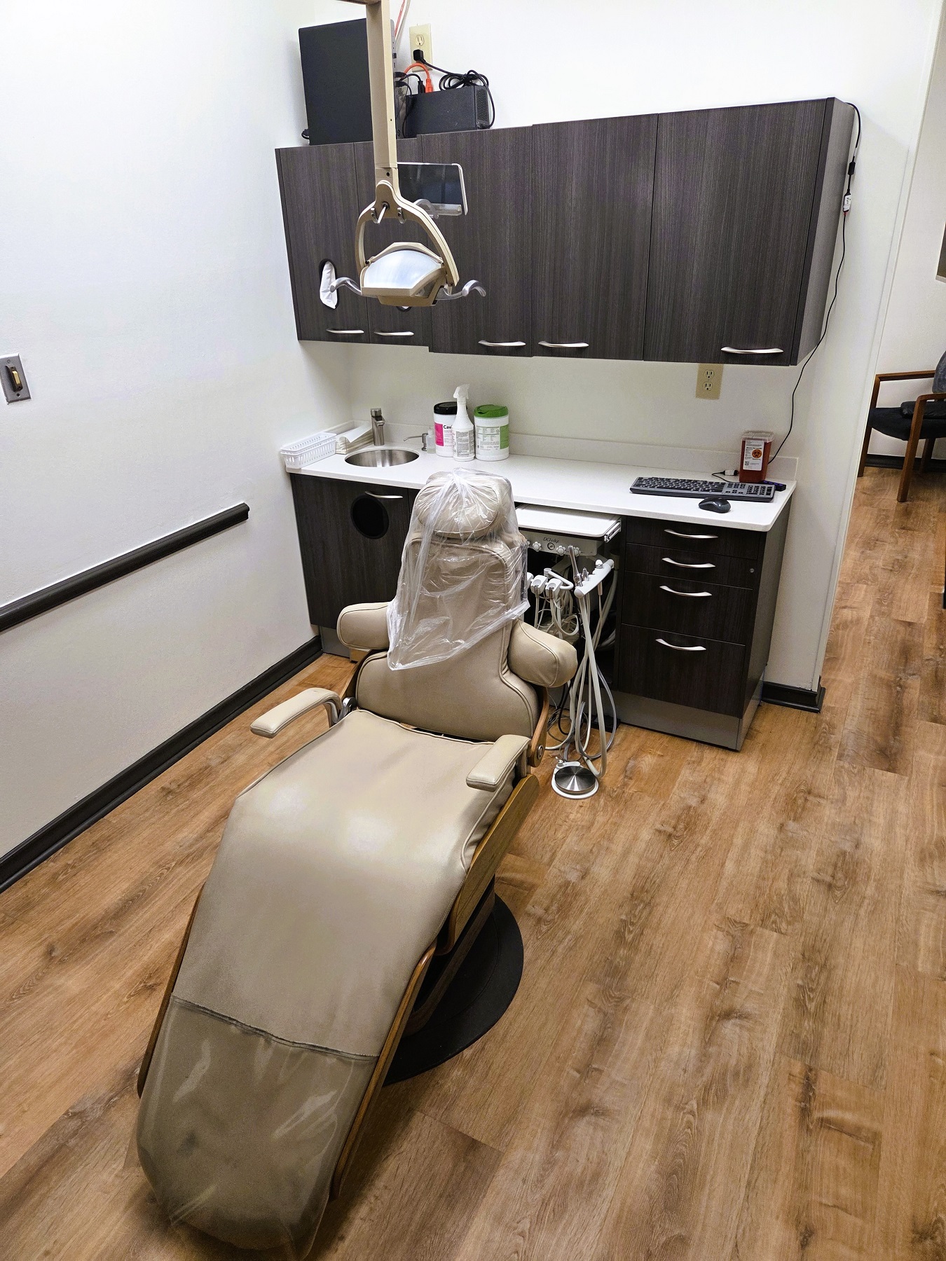 Light walls with dark stained baseboards and chair rail and medium stained wood flooring. A cushioned dental procedural chair anchors the room with an overhead dental light. A cabinet with sink, shelves hold lots of equipment, tools, and supplies.